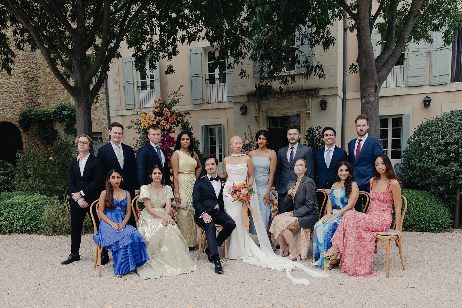 Group picture of guests dressed in formal attire at the end of the wedding ceremony outdoor, with trees and Chateau La Tour Vaucros with shutters in the background, celebrating a special occasion like a wedding.