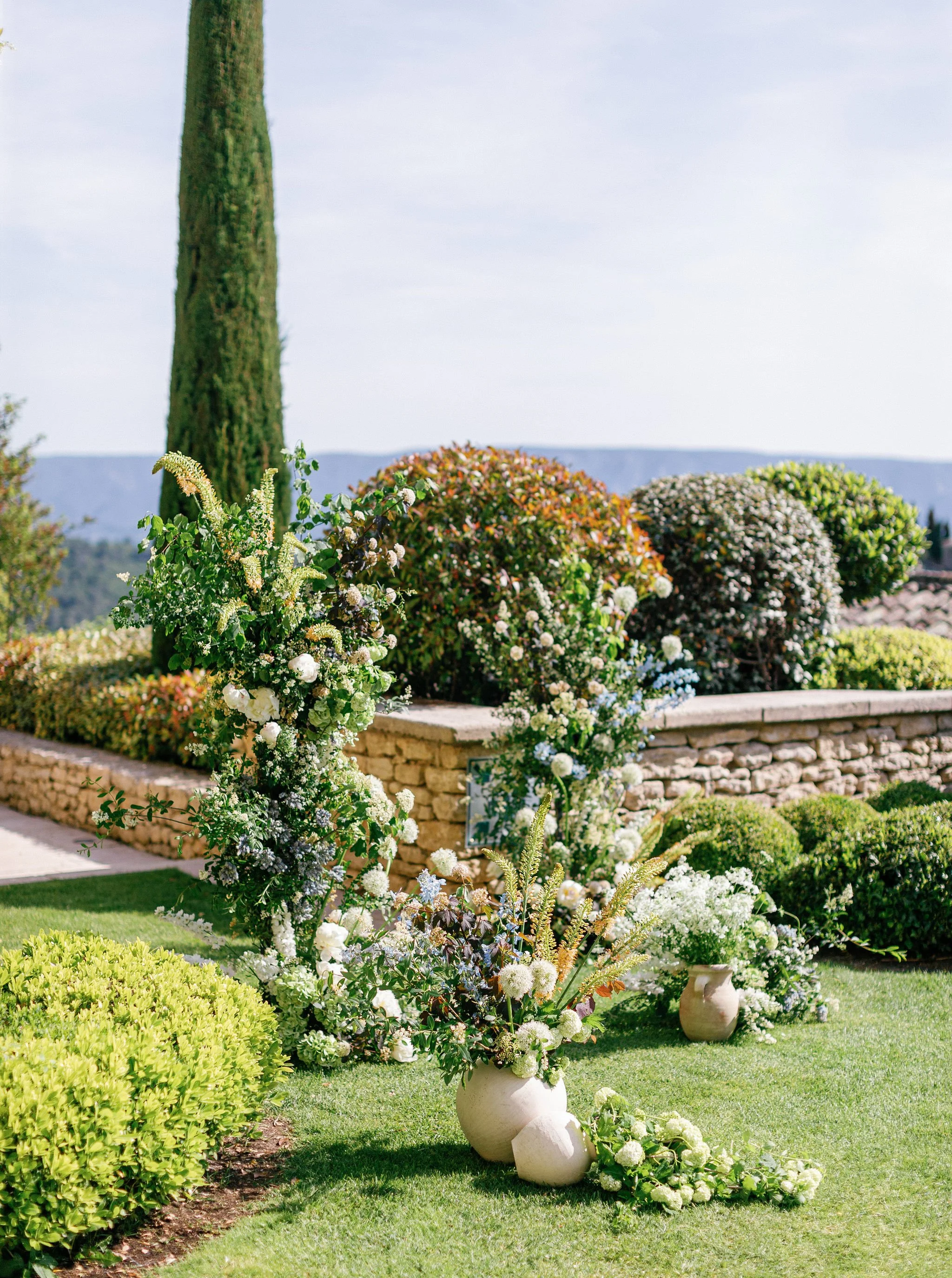 An asymetrical arch in Bastide de Gordes' garden with large vases filled with white and green floral arrangements, surrounded by trimmed bushes and a stone wall, under a clear sky.