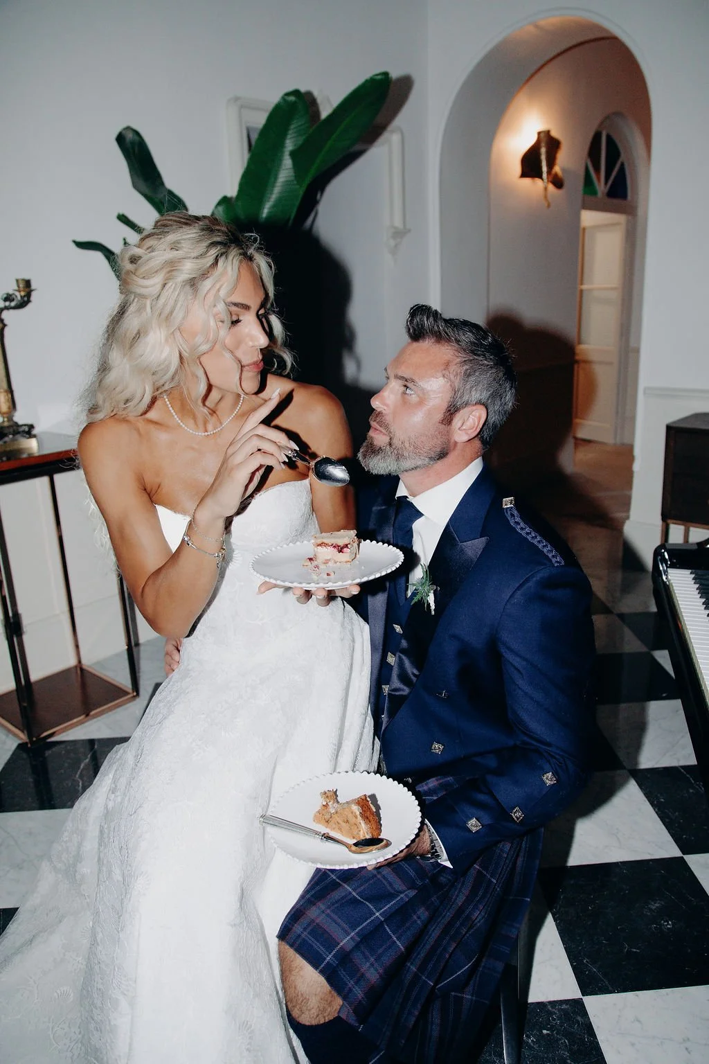The bride feeds a slice of cake to her groom in a kilt, who is seated and holding a plate with another slice of cake.