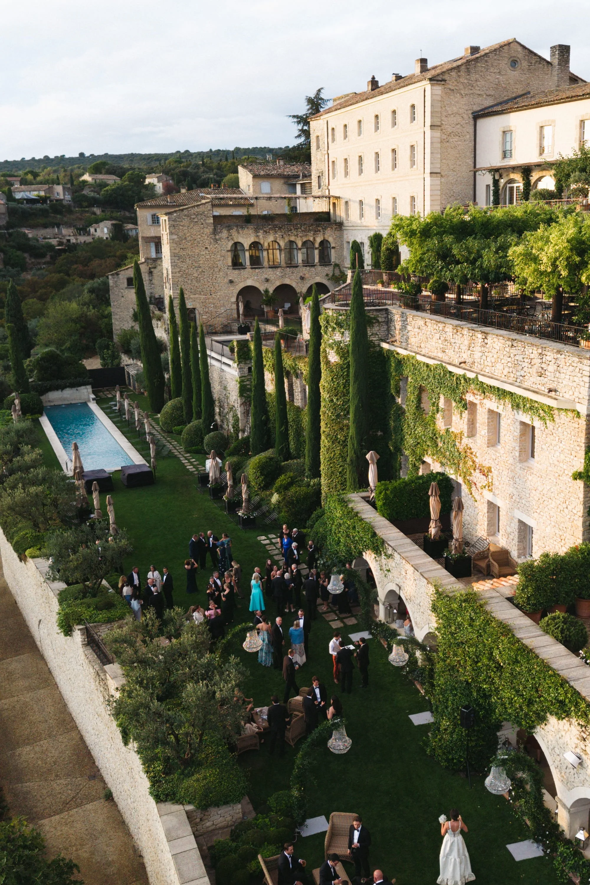 Wedding reception outdoor on a lush green terrace with a long pool, surrounded by tall cypress trees and stone buildings, with guests mingling and a bride in a white dress. At La Bastide de Gordes in Provence.