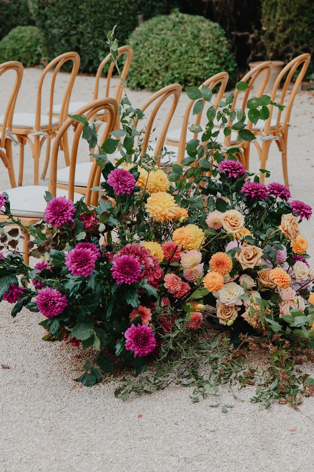 A floral arrangement with pink, yellow, and cream roses and dahlias, set on the ground in front of a row of wooden chairs with white cushions, outdoors on sandy ground with green bushes in the background.
