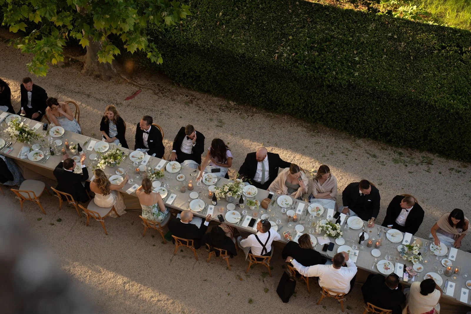 Aerial view of a long formal dinner table outdoors with guests dressed in formal attire, including suits and dresses, dining and celebrating.