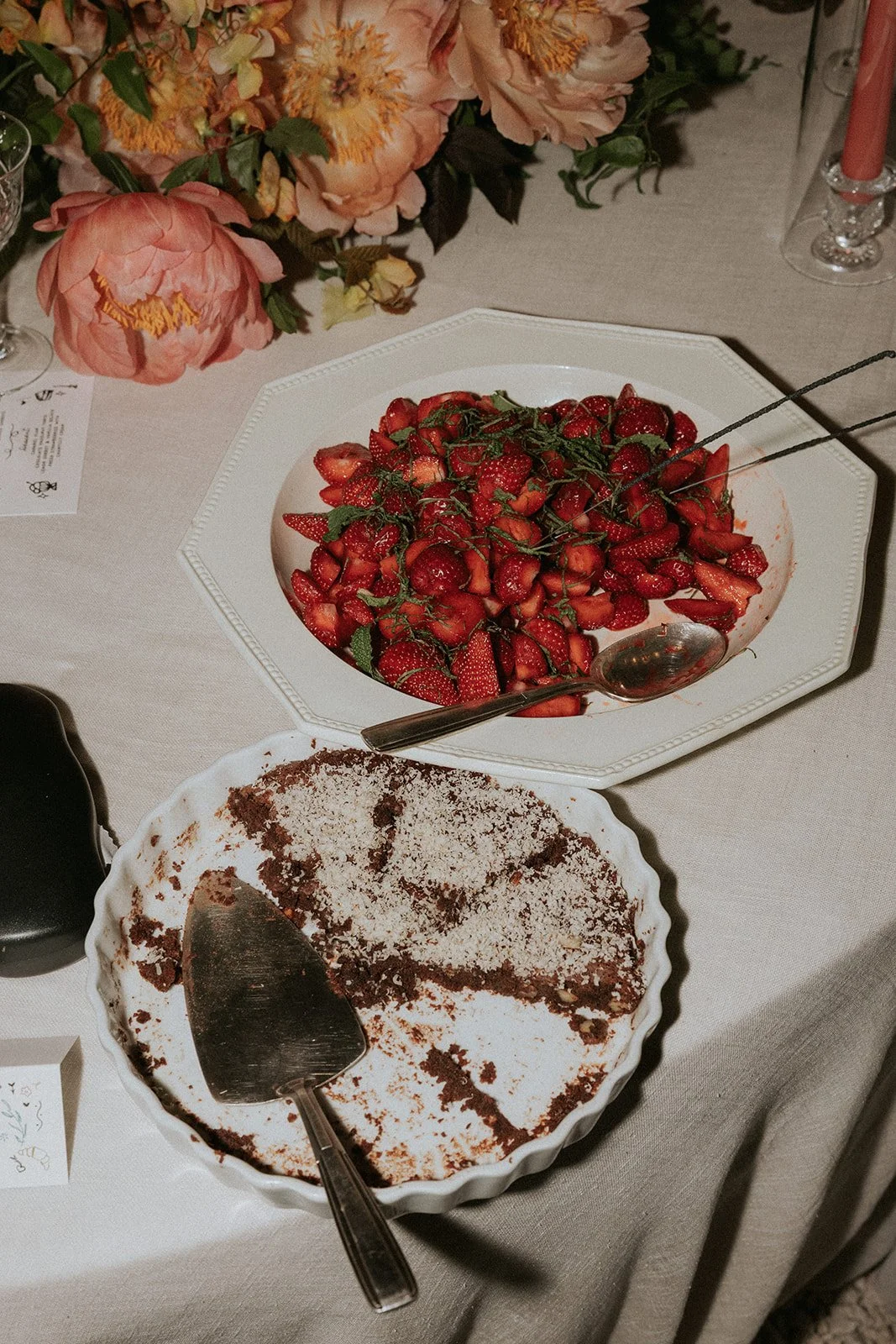 A table with a large plate of sliced strawberries, a serving spoon, and a partially eaten chocolate dessert in a white dish, with pink flowers in the background.