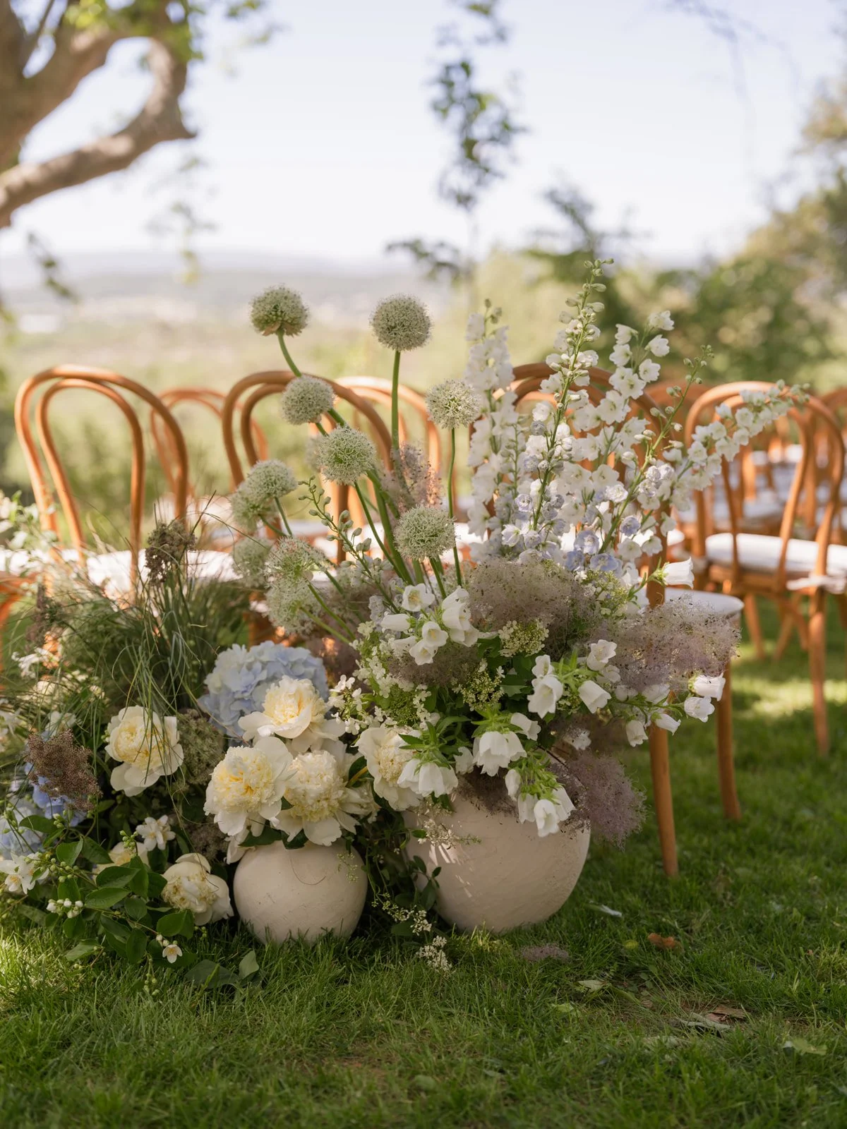 Decorative flower arrangement with white and light blue flowers in white vases, set outdoors with wooden chairs in the background.