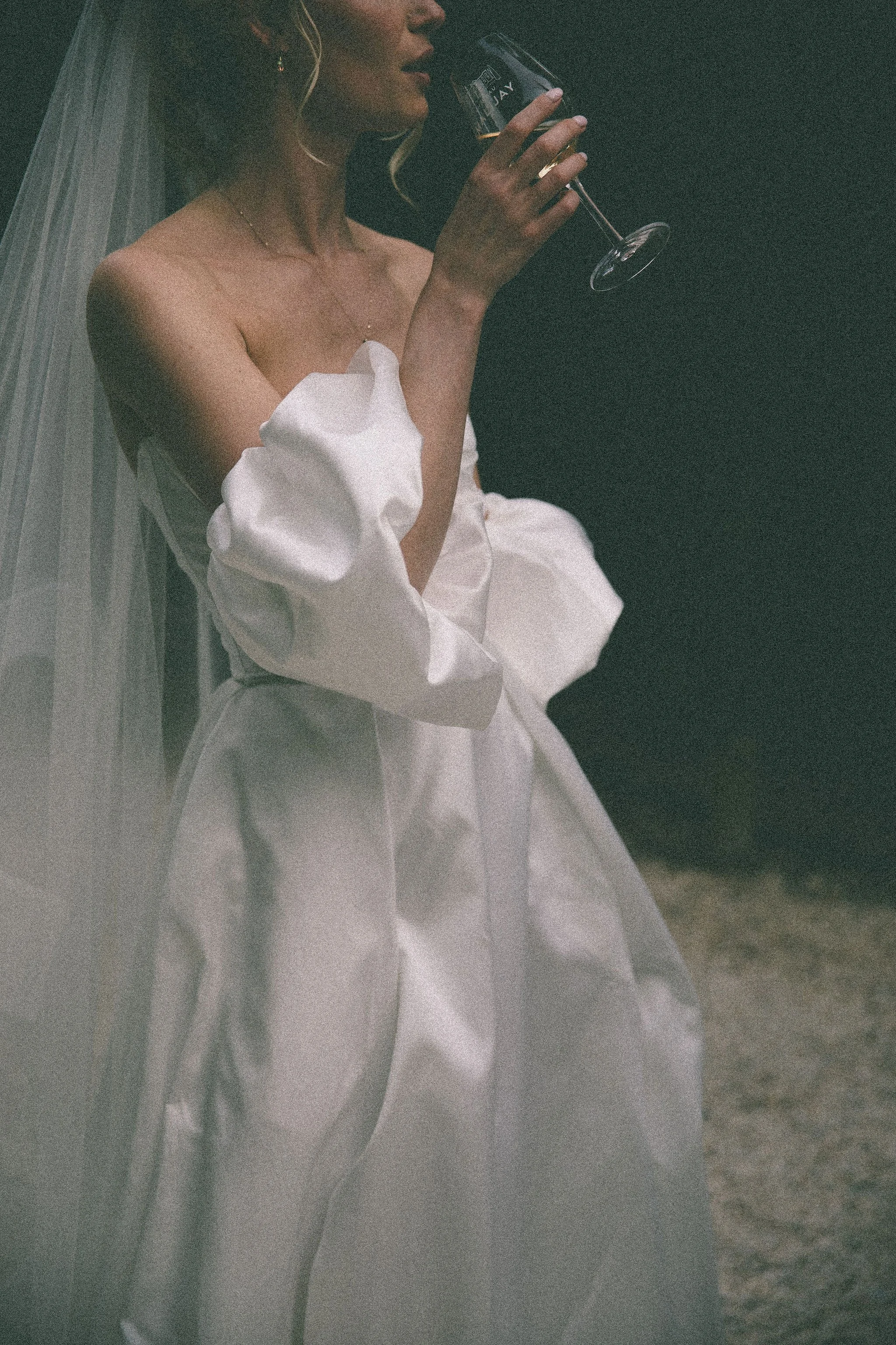 A woman in a white dress and veil, holding a glass of dark beverage, standing in a dark environment.