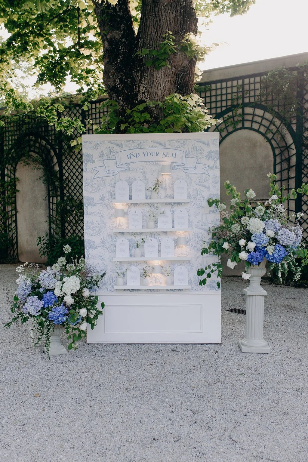 A wedding seating chart display with a white background, floral decorations, and shelves with cards. Large floral arrangements in white and blue vases flank the display, set outdoors under a large tree with sunlight filtering through the leaves.