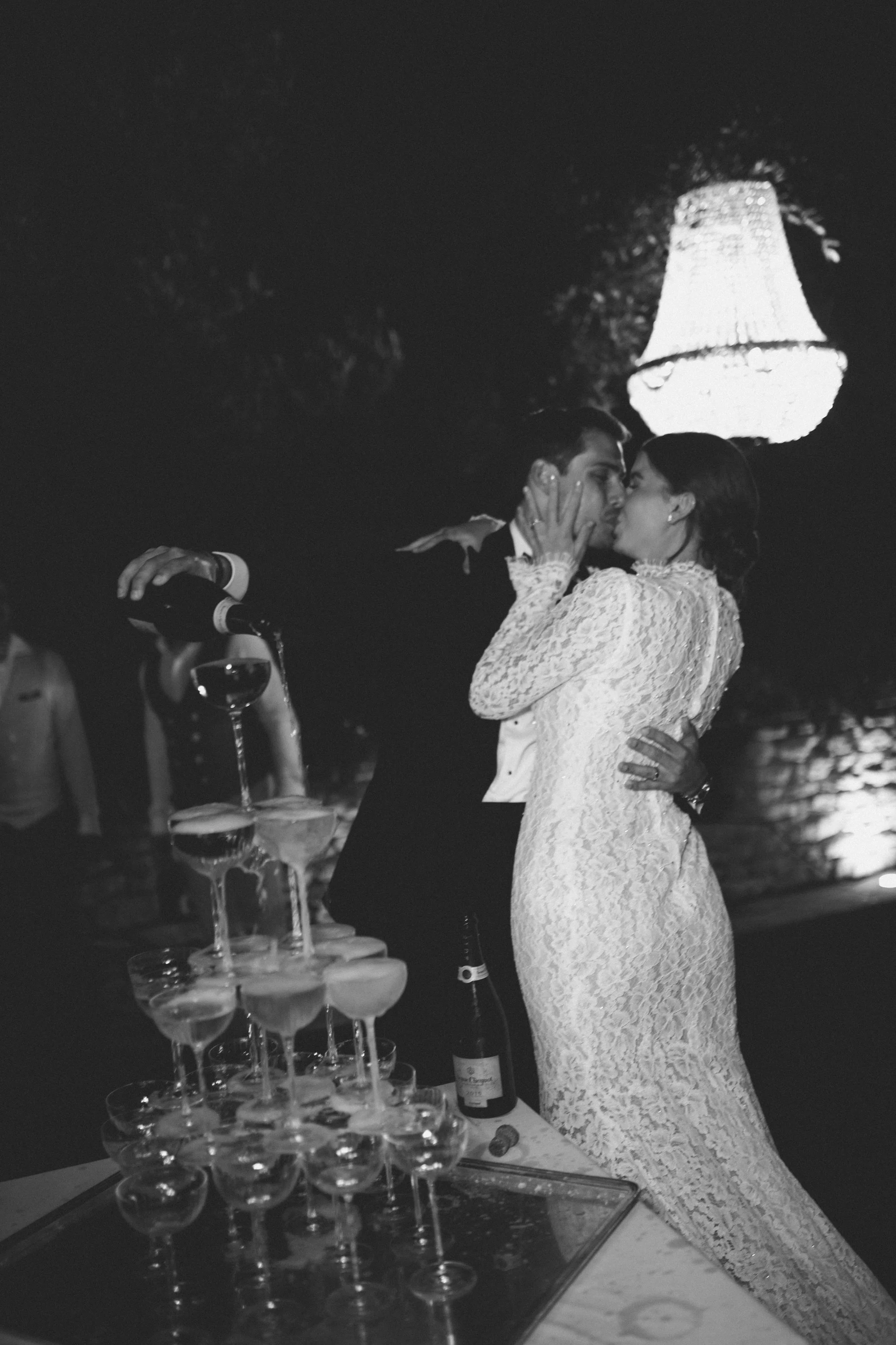 A black and white photo of a bride and groom kissing during their wedding reception, at the champagen tower, with a large chandelier hanging above them.