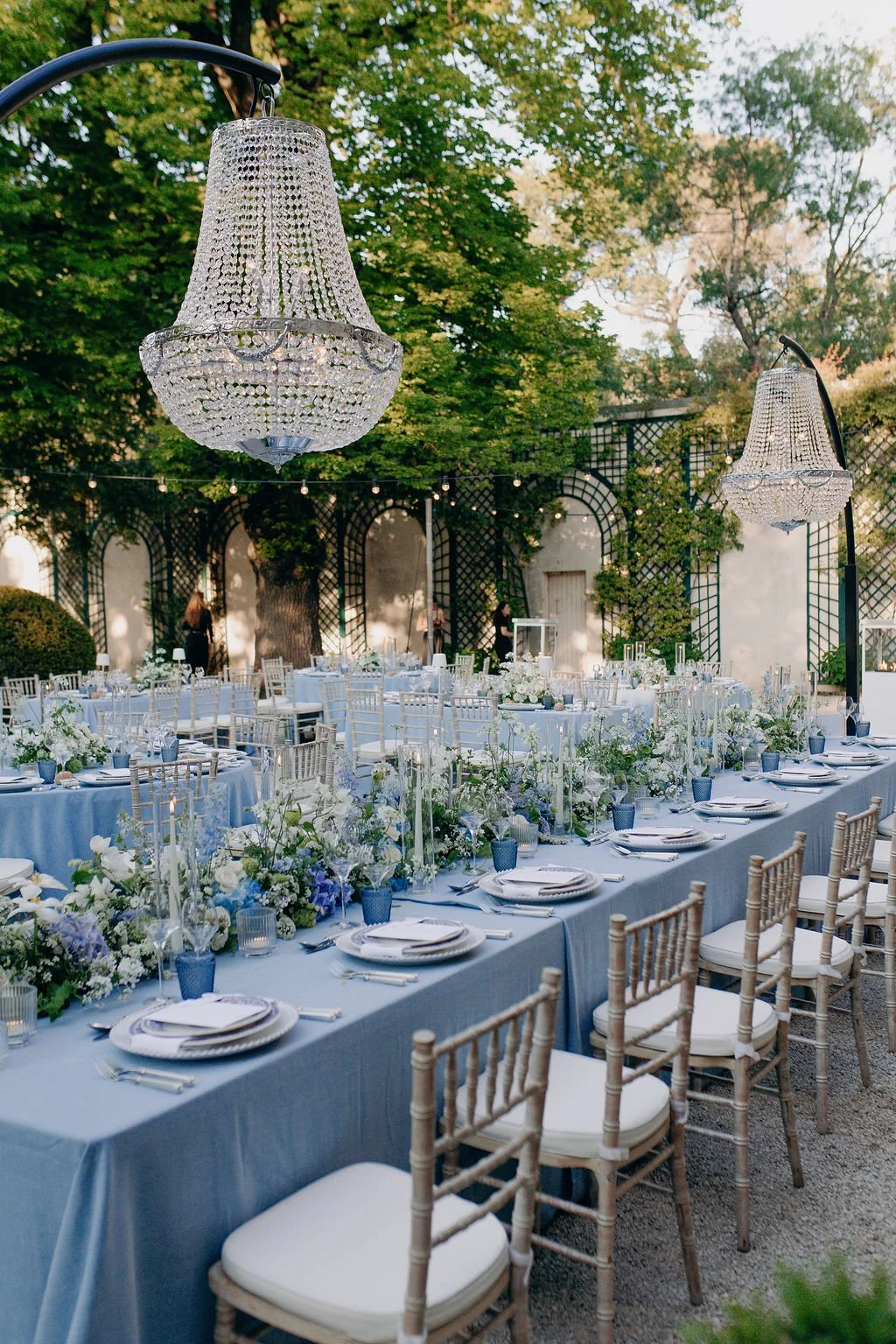 Elegant outdoor wedding reception with long tables draped in light blue tablecloths, decorated with white flowers and greenery, and illuminated by two large crystal chandeliers hanging overhead.