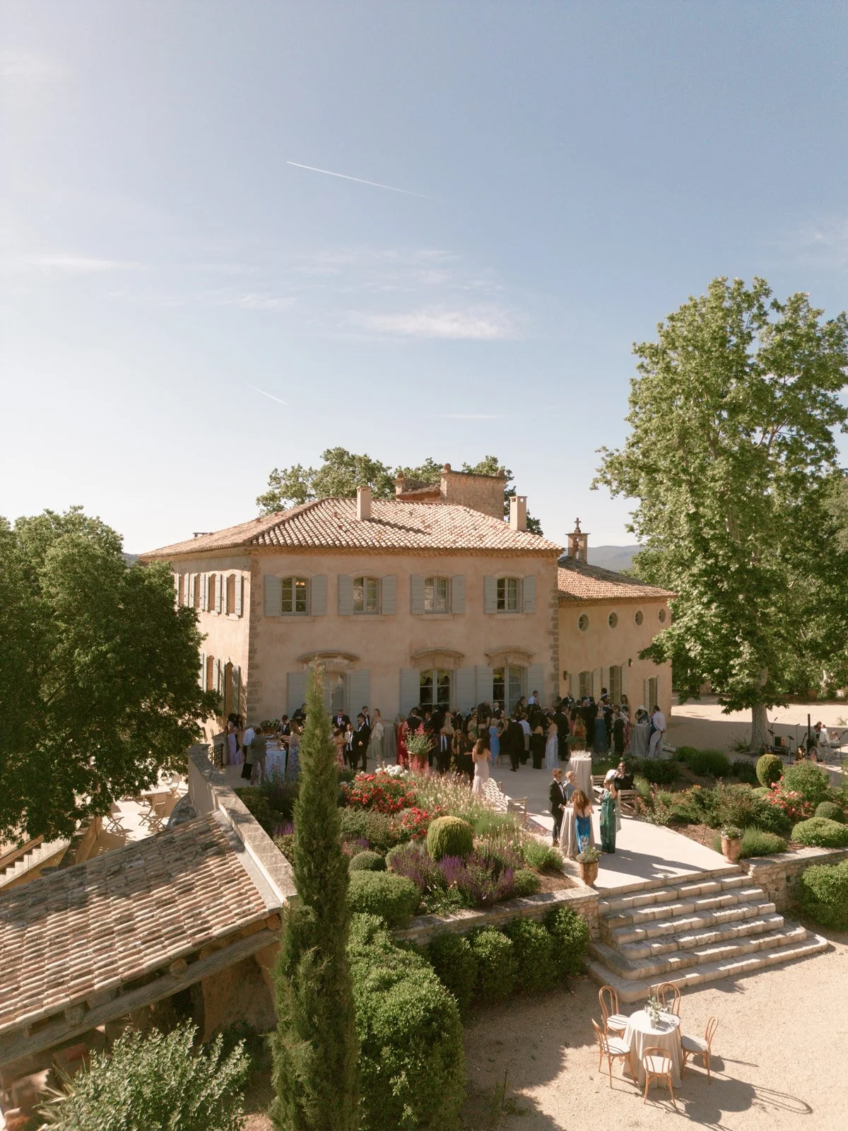Guests gather outside cocktail hour in Provence, elegant mansion with a tiled roof and multiple windows during a celebration, with steps leading up to the entrance, surrounded by well-maintained gardens and trees on a sunny day.