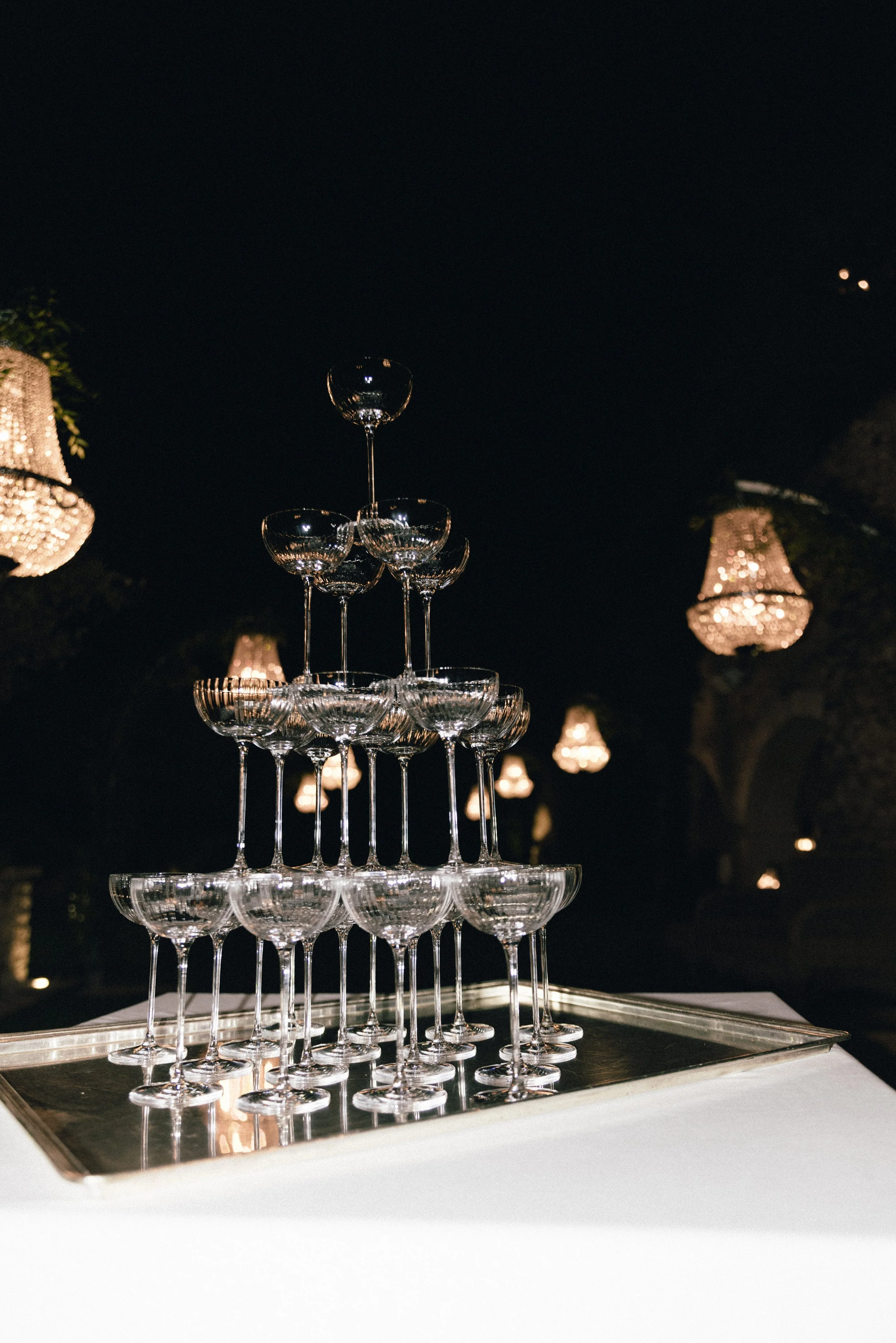 A pyramid of stacked empty champagne glasses on a tray at night, champagne tower, decorated with hanging chandeliers in the background.