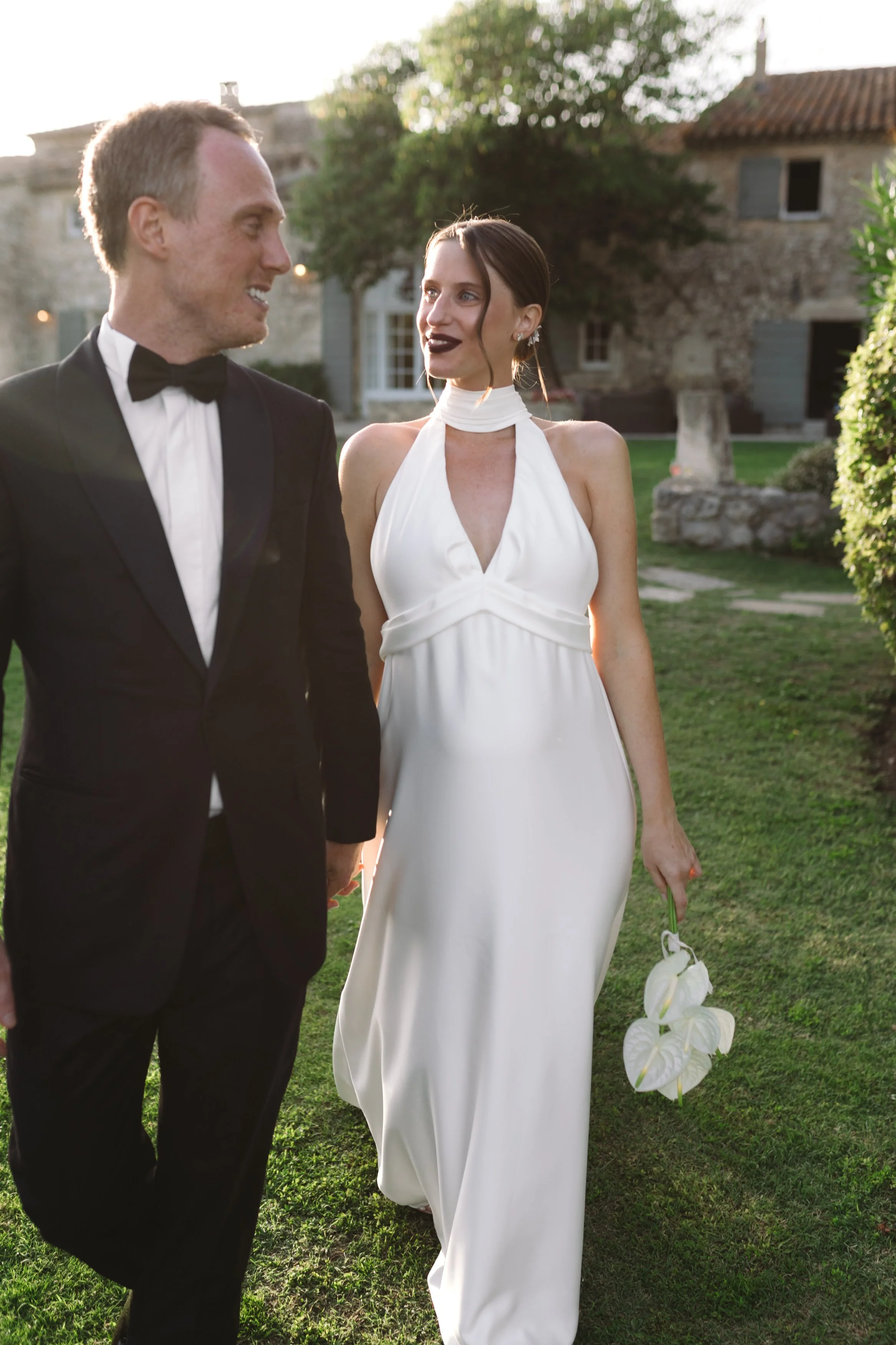 A couple dressed in formal attire, the man in a tuxedo and the woman in a white dress, holding a small bouquet, walking outdoors in a garden area during sunset, with Mas de la Rose in the background.