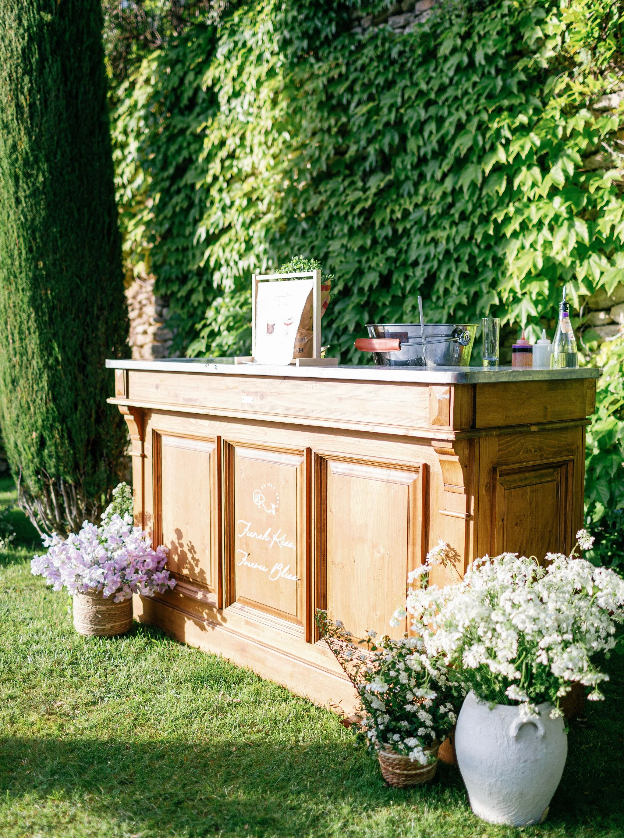 Wooden beverage bar setup outdoors with flowers, greenery, and drink supplies.