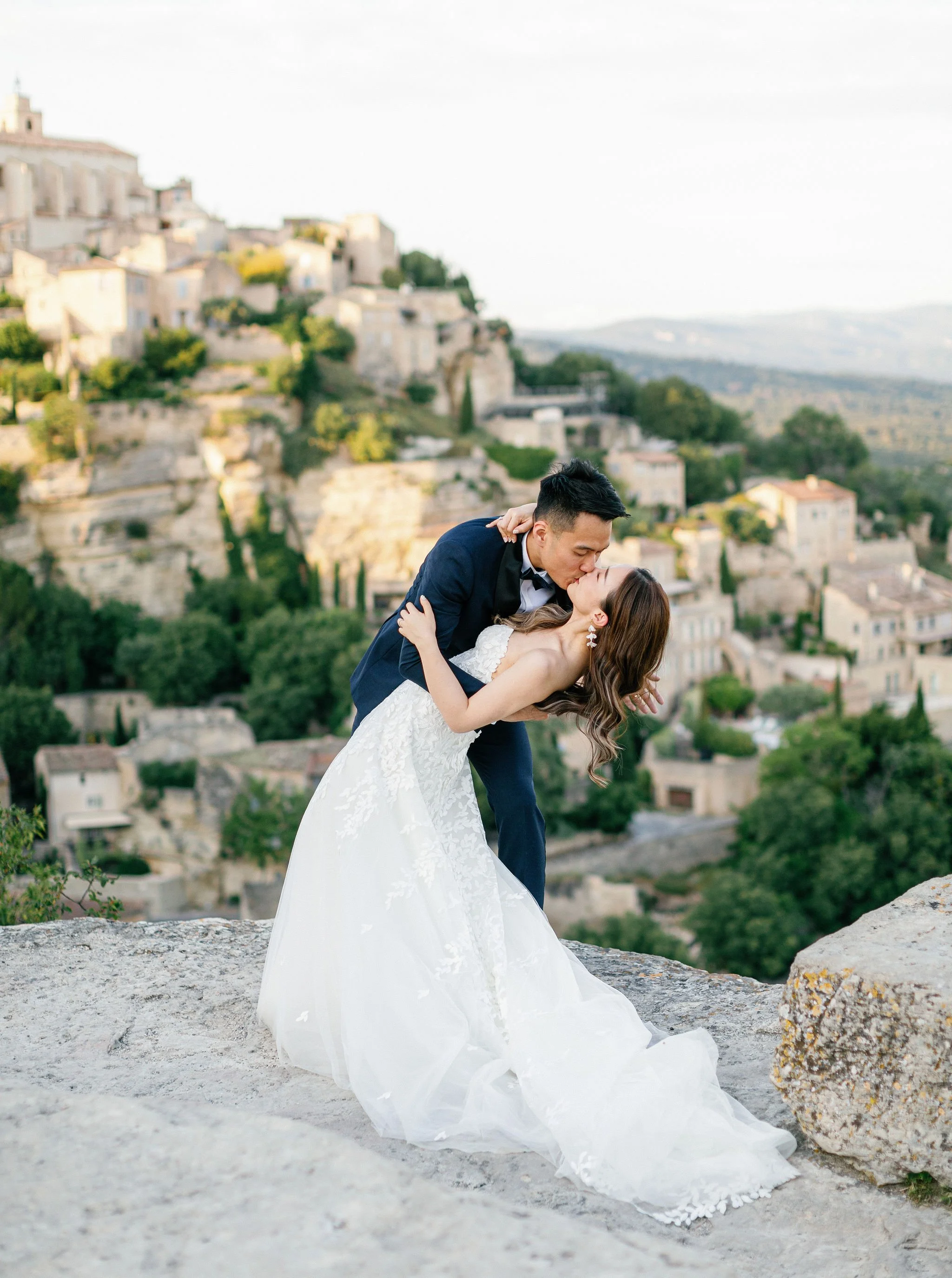 A newlywed couple shares a kiss on a rocky ledge with Gordes village in the background.
