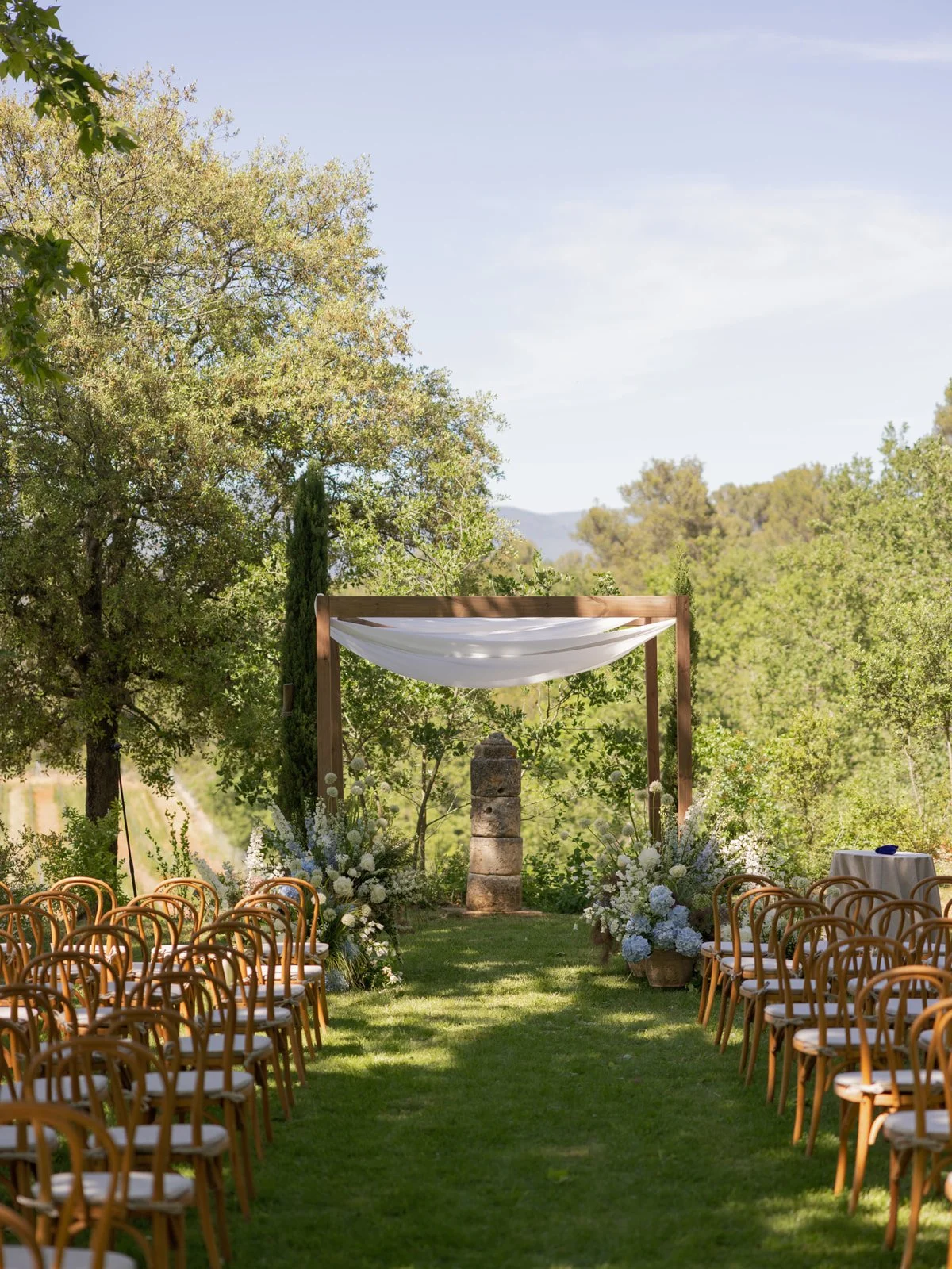 Outdoor wedding ceremony setup with wooden chairs on either side of a grassy aisle, floral arrangements, and a wooden arch with white fabric draping, surrounded by trees under a clear blue sky.