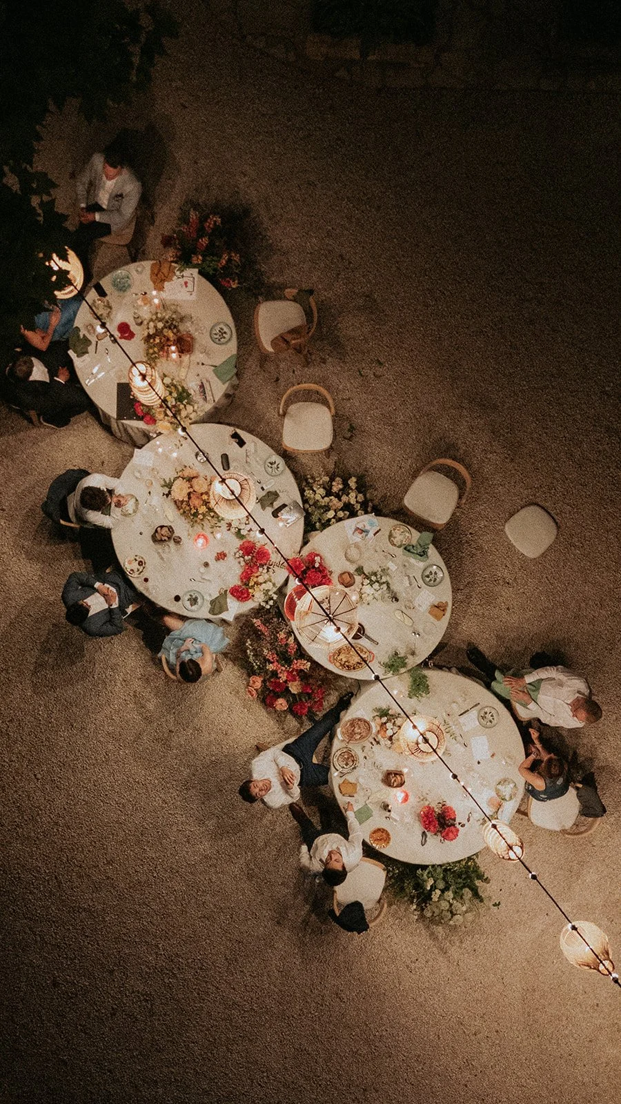 Overhead view of a wedding dinner party with four round tables decorated with flowers and candles, surrounded by guests in formal attire, set on a gravel surface illuminated by hanging string lights. Micro wedding.