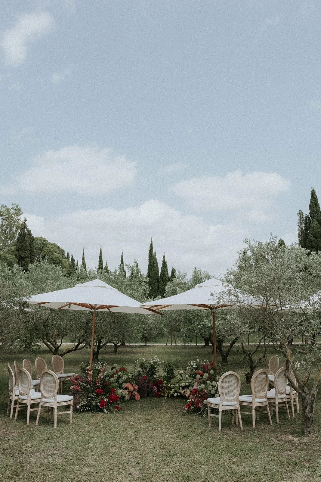 Outdoor wedding ceremony setup with white umbrellas, floral arrangements, wooden chairs, and trees under a partly cloudy sky.