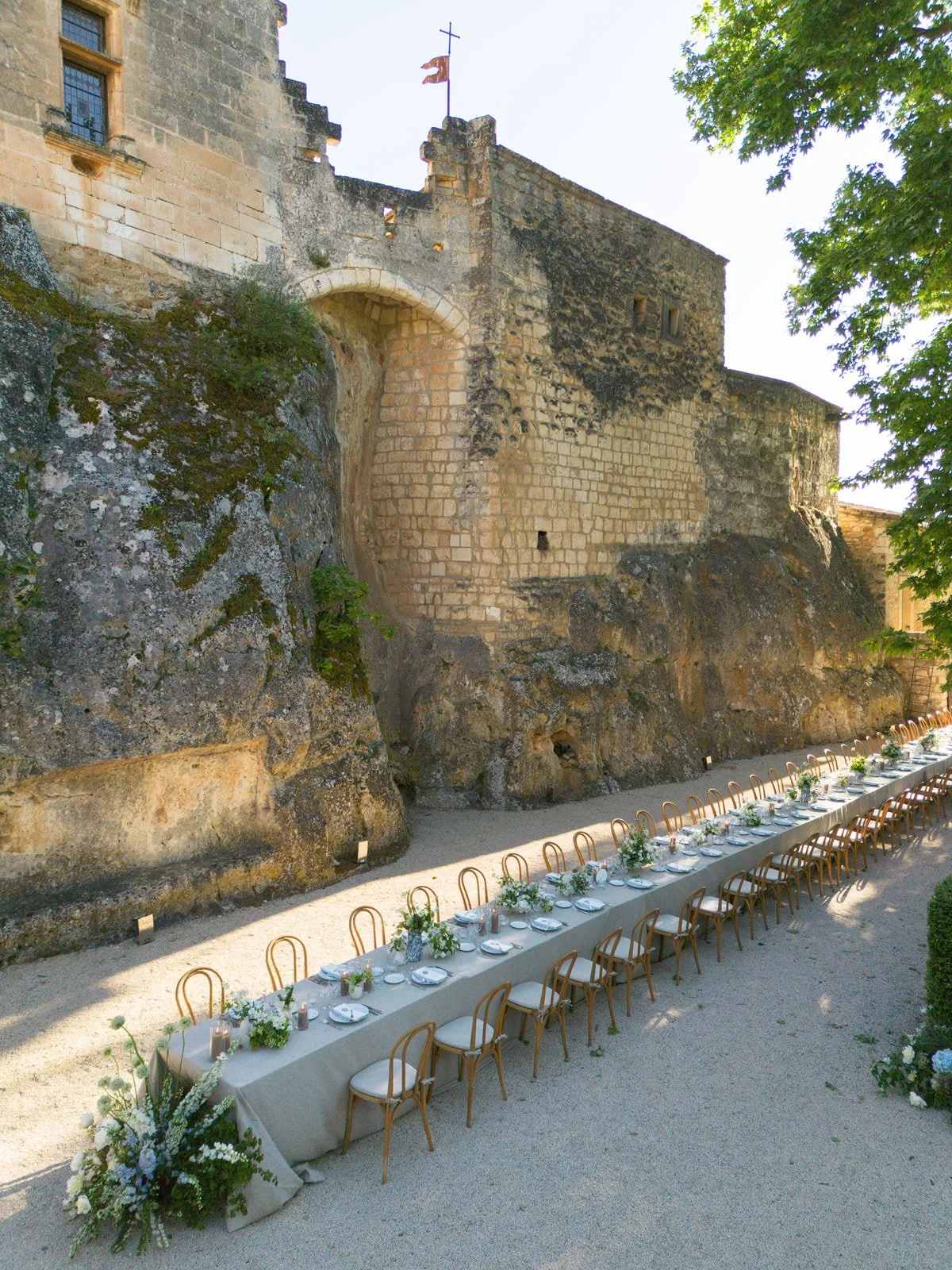 Outdoor banquet setup with a long table decorated with white tablecloth, flowers, and place settings, situated against the base of an ancient stone castle wall.