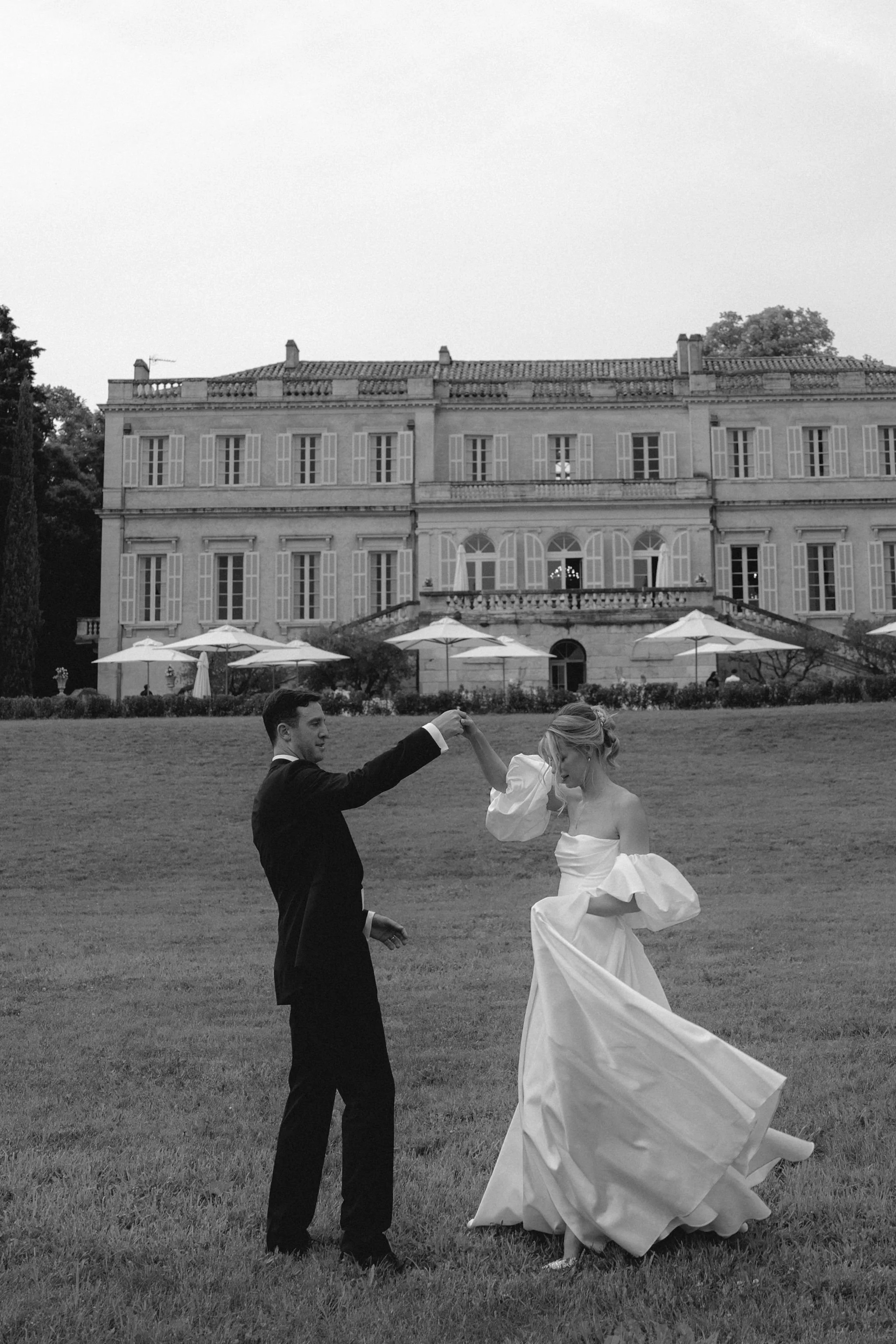 A black-and-white photo of a bride and groom dancing outside a large estate or mansion. The groom is wearing a suit and the bride is in a wedding dress, holding the skirt as they dance on the grass in front of CHateau Martinay, Provence.