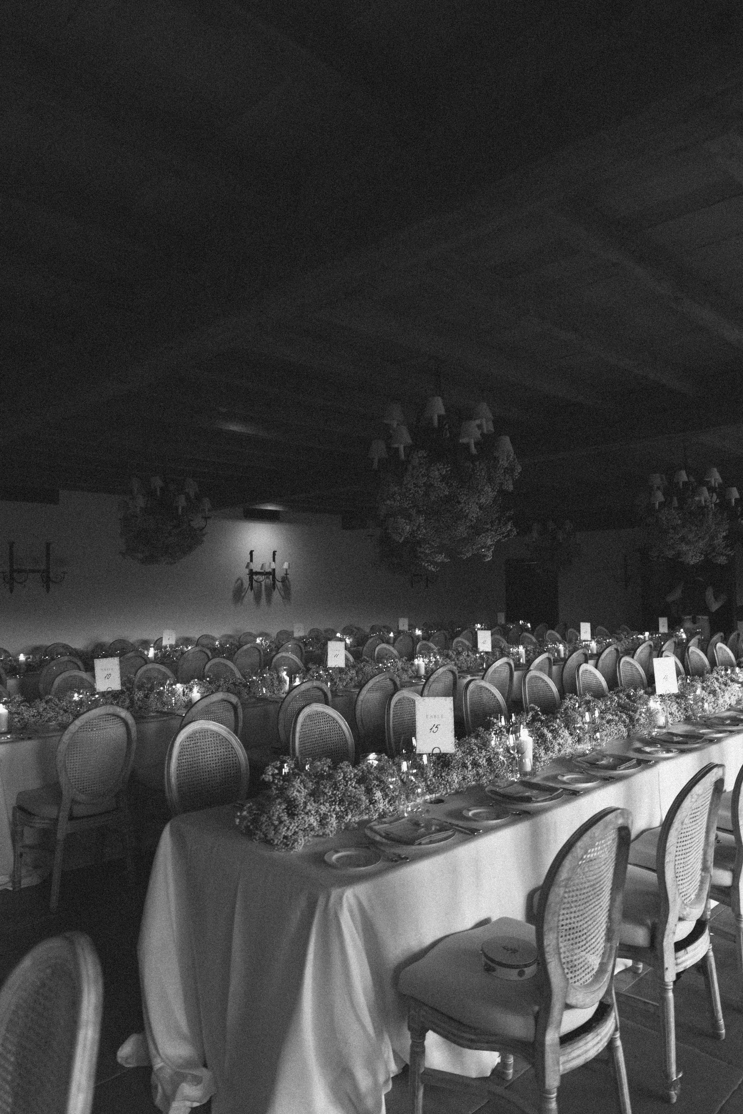 A large banquet hall decorated for an event with long tables adorned with baby's breath centerpieces, candles, and place settings, chairs arranged around the tables, and chandeliers hanging from a wooden ceiling.