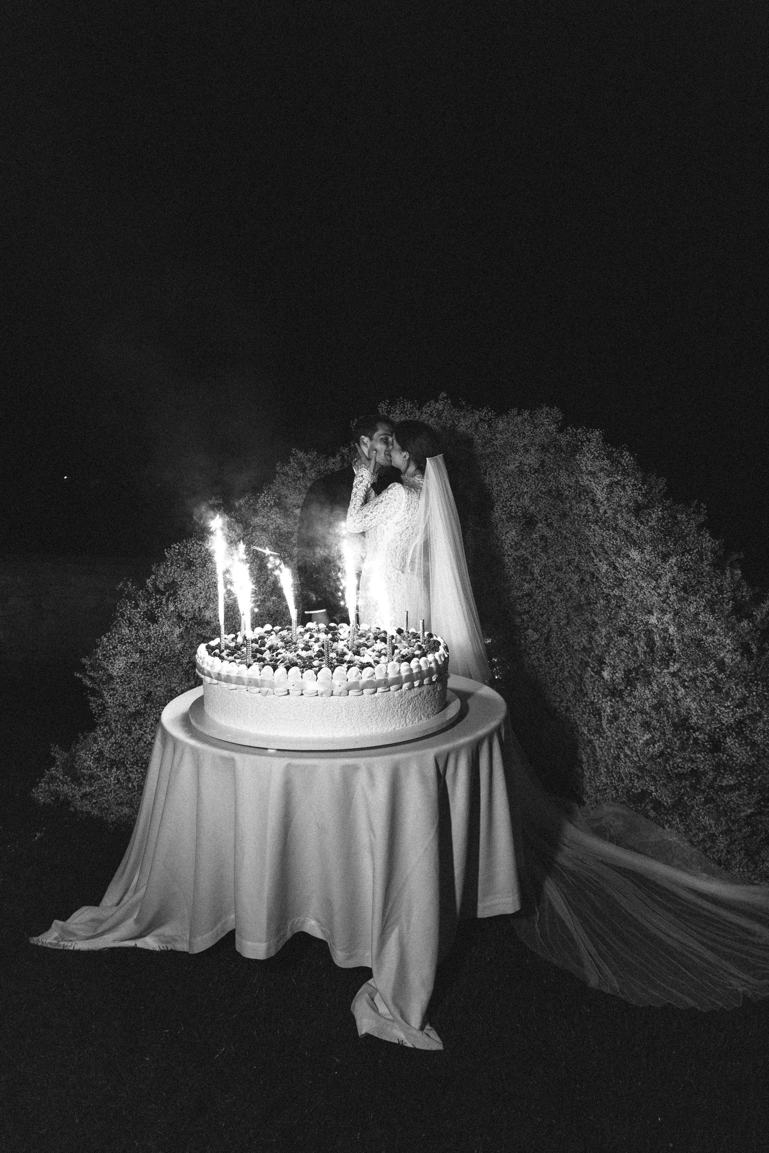 A bride and groom sharing a kiss at night during their wedding celebration, with a giant pavlova as the wedding cake on a table in front of them, decorated with sparklers and surrounded by huge baby's breath arch.