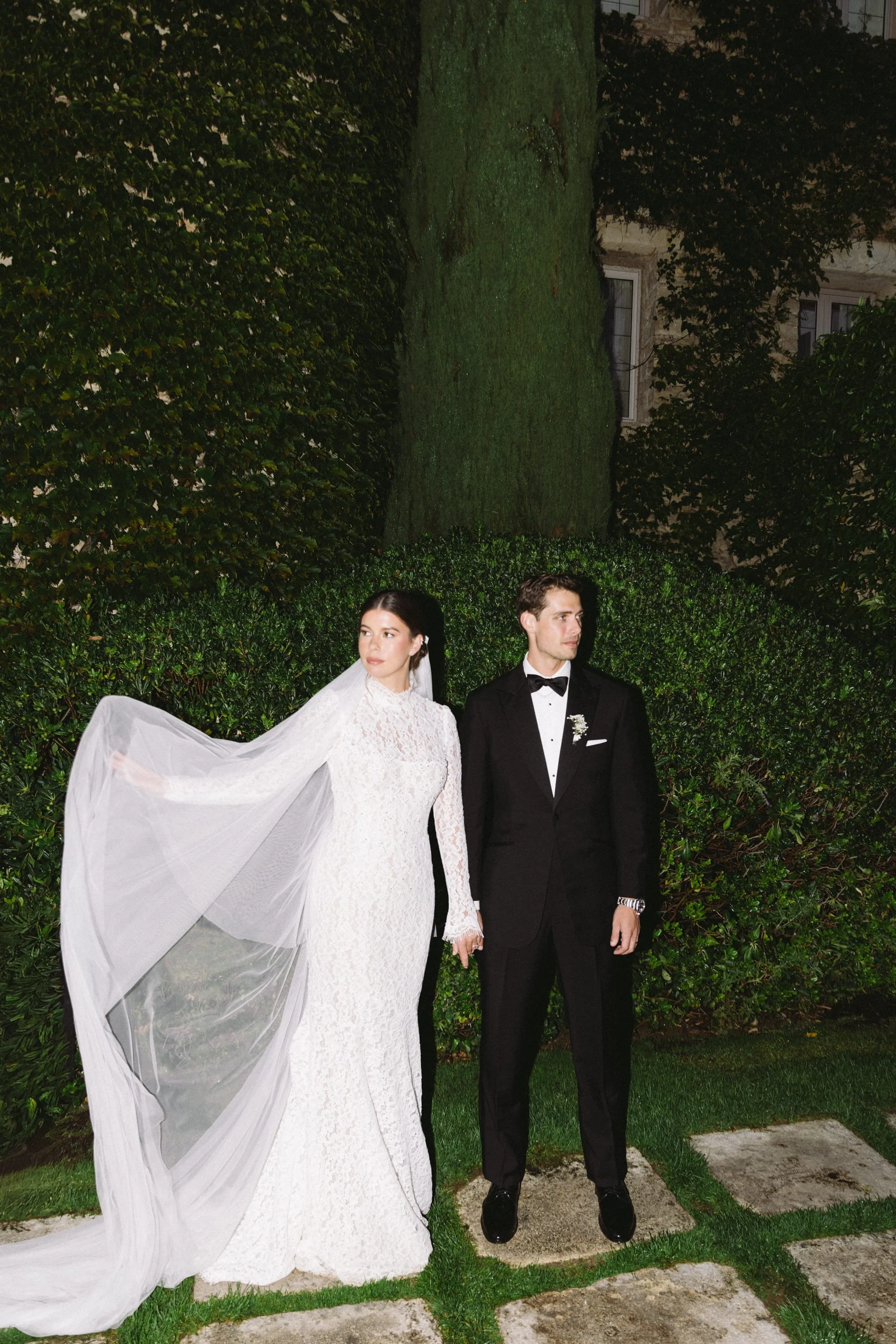 Bride and groom standing outdoors in wedding attire, holding hands, with a green hedge and trees in the background. Couple session.