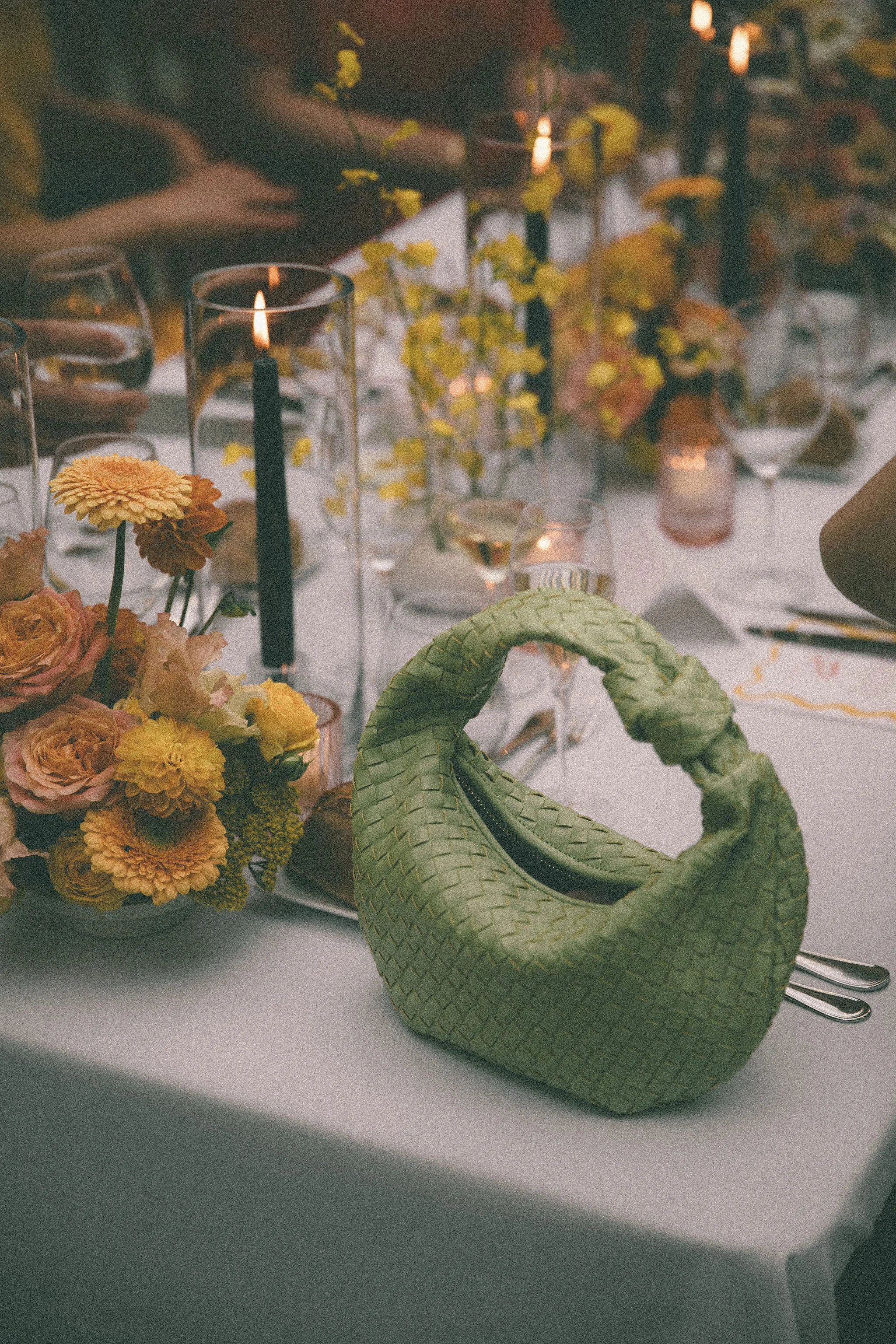 A green woven handbag on a table decorated with a floral centerpiece, candles, and glasses at an elegant wedding dinner.