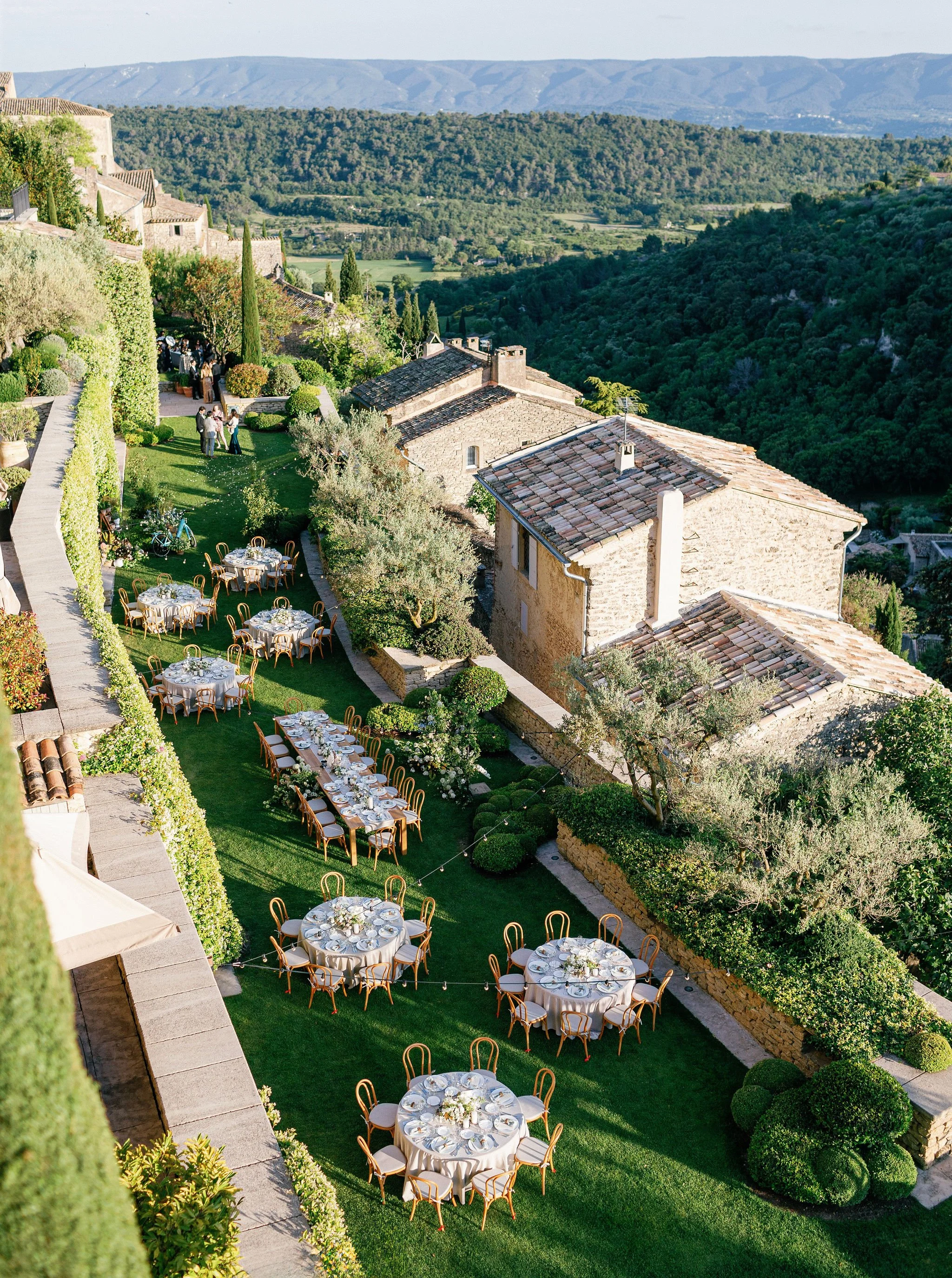 Outdoor wedding on a lush green lawn of La Bastide de Gordes in Provence with round dining tables adorned with white tablecloths and floral centerpieces, surrounded by wooden chairs.
