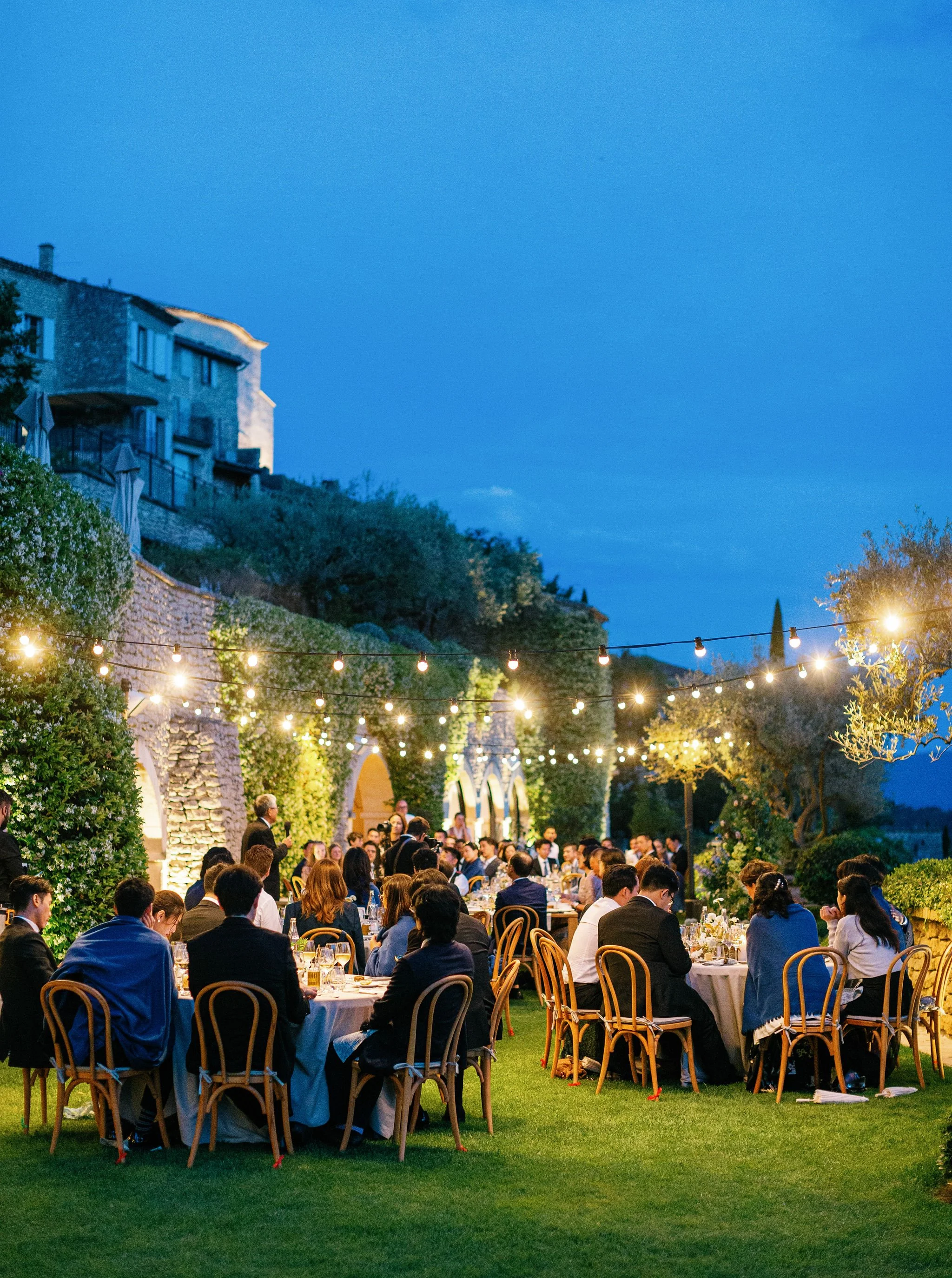 A large outdoor gathering with numerous guests seated at round tables, decorated with white tablecloths and floral centerpieces. The event takes place in a lush garden space under string lights at dusk, with a stone building and hillside in the backg