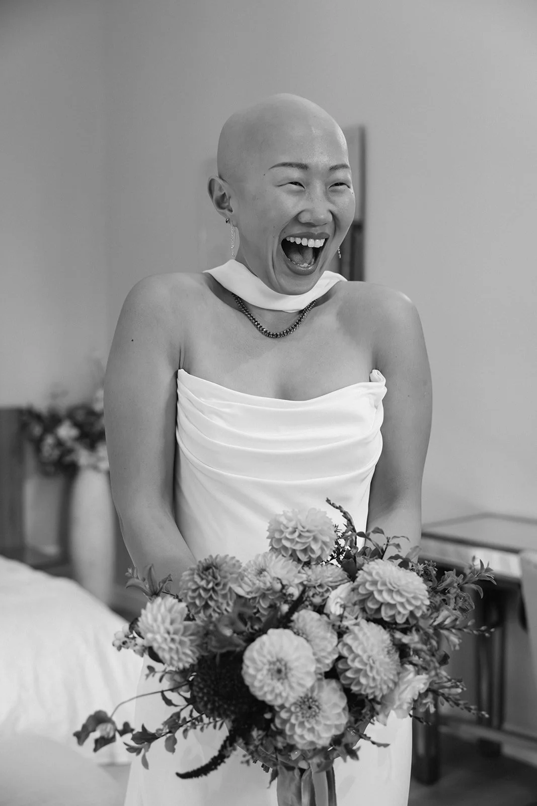Black and white photo of a woman in a wedding dress, holding a bouquet of flowers, and laughing joyfully.