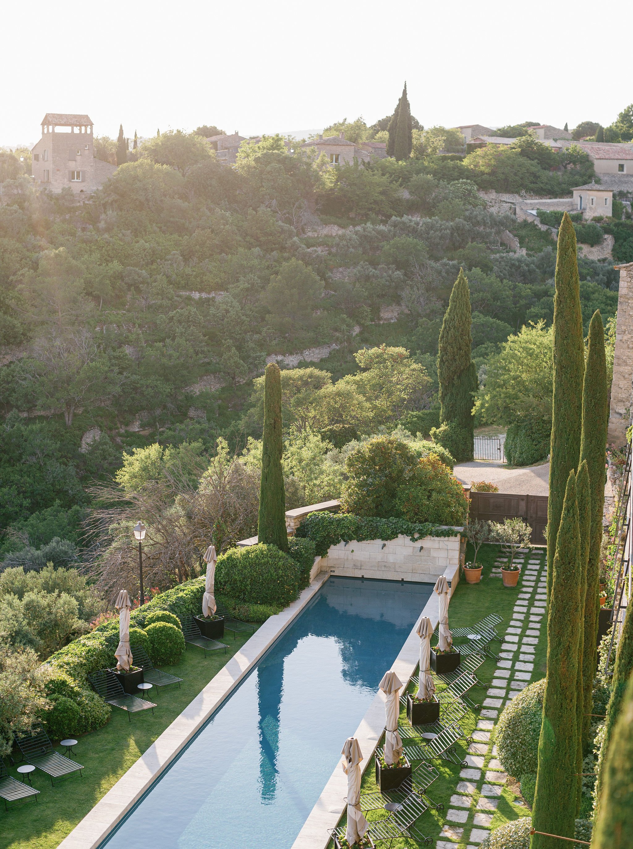 Photograph of a luxurious backyard with a swimming pool of La Bastide de Gordes in Provence, surrounded by tall cypress trees and greenery, with a view of a hilly landscape and houses in the background.