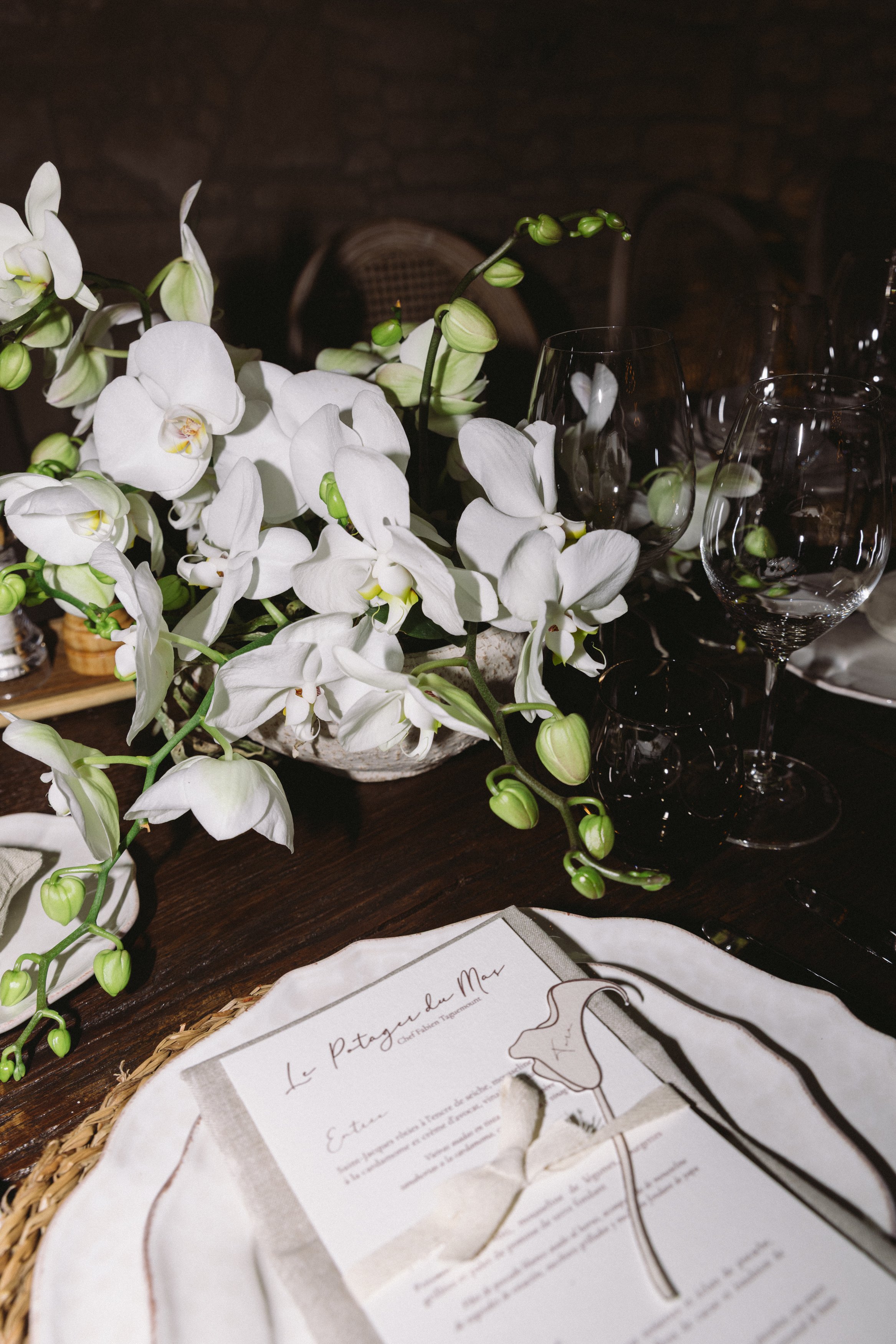 A table decorated with a bouquet of white orchids and glassware, with a wine menu on a placemat in the foreground.