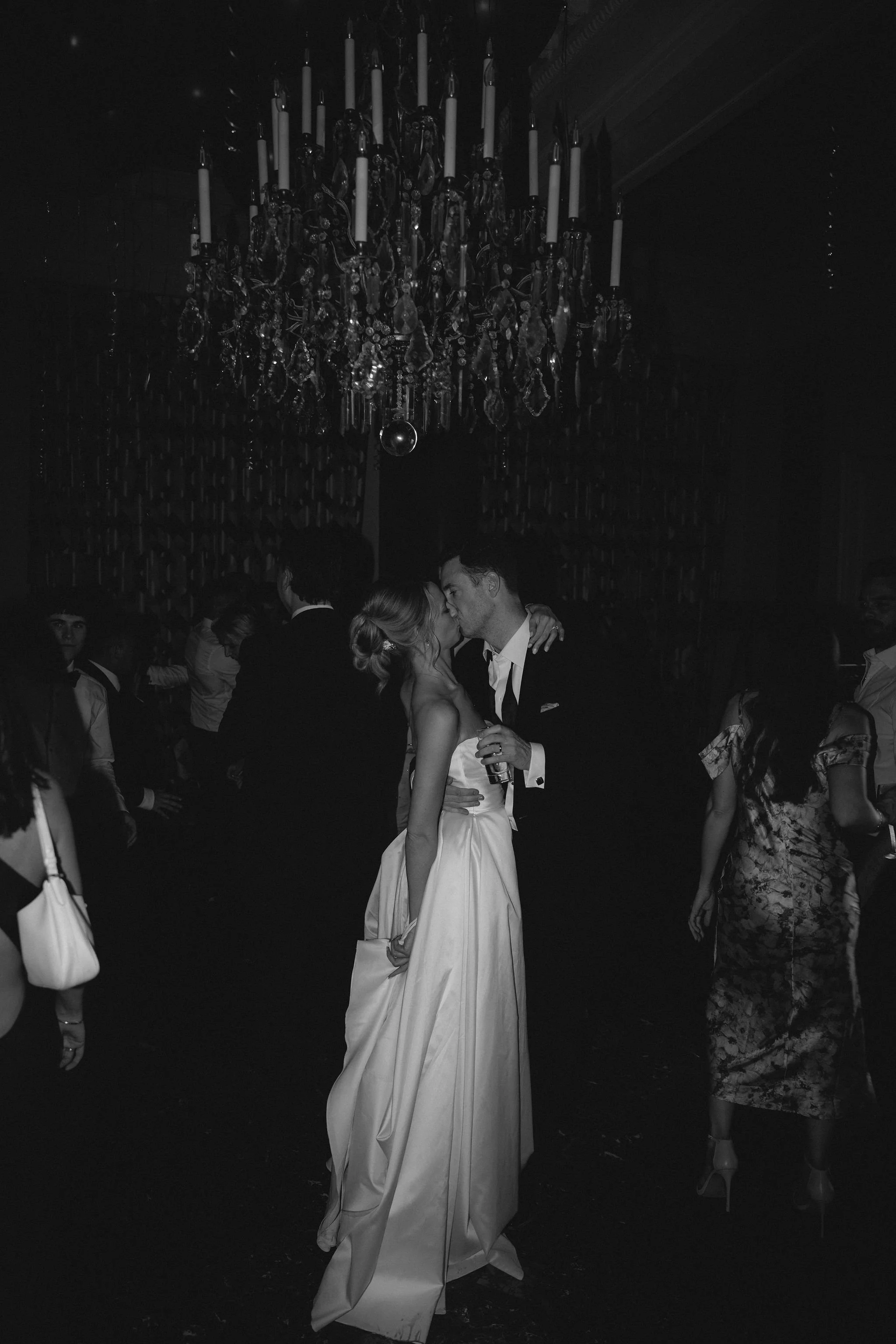 A couple in wedding attire kissing at a dimly lit party with a chandelier hanging overhead and other guests in the background.