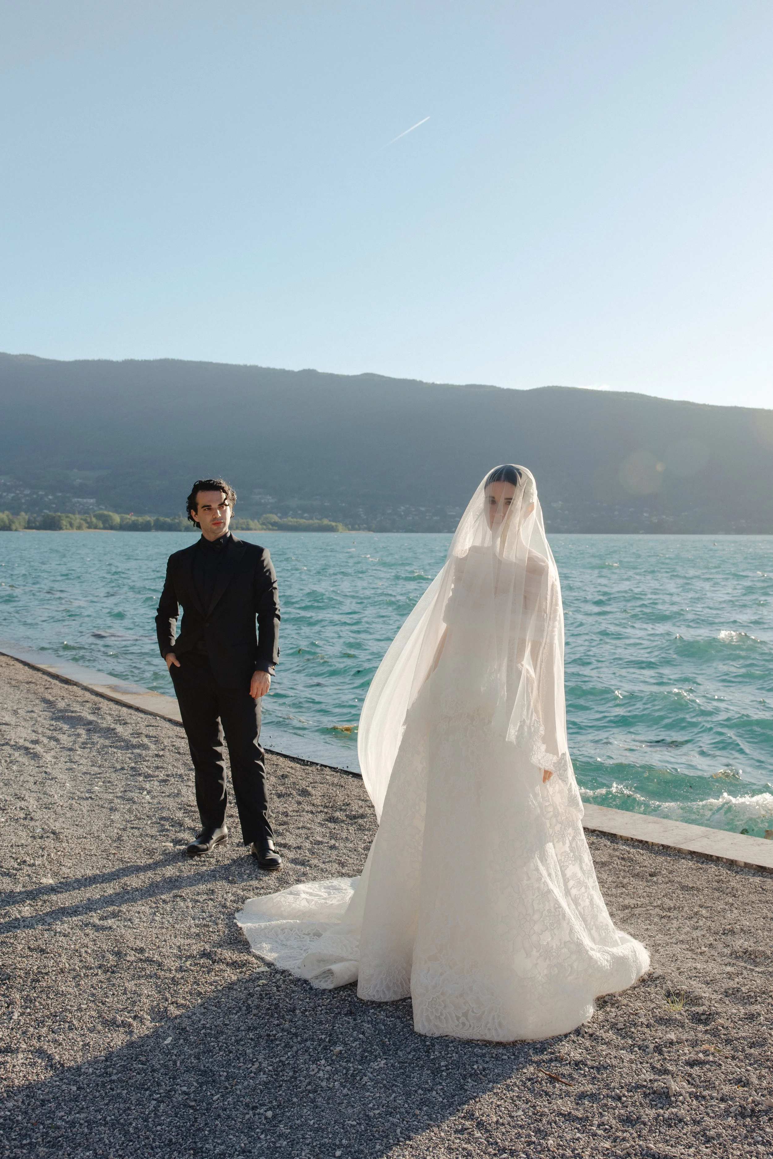 A bride in a white wedding dress and veil standing on a rocky lakeside with a groom in a black suit nearby, mountains in the background, and water with small waves.