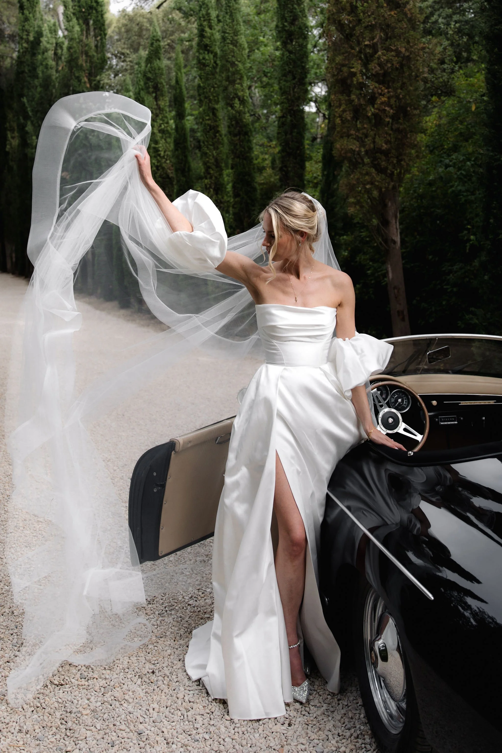 Bride in a white wedding dress with a slit, sitting on a vintage black car with a beige interior, holding a flowing veil outdoors surrounded by cypress at Charteau Martinay, Provence.