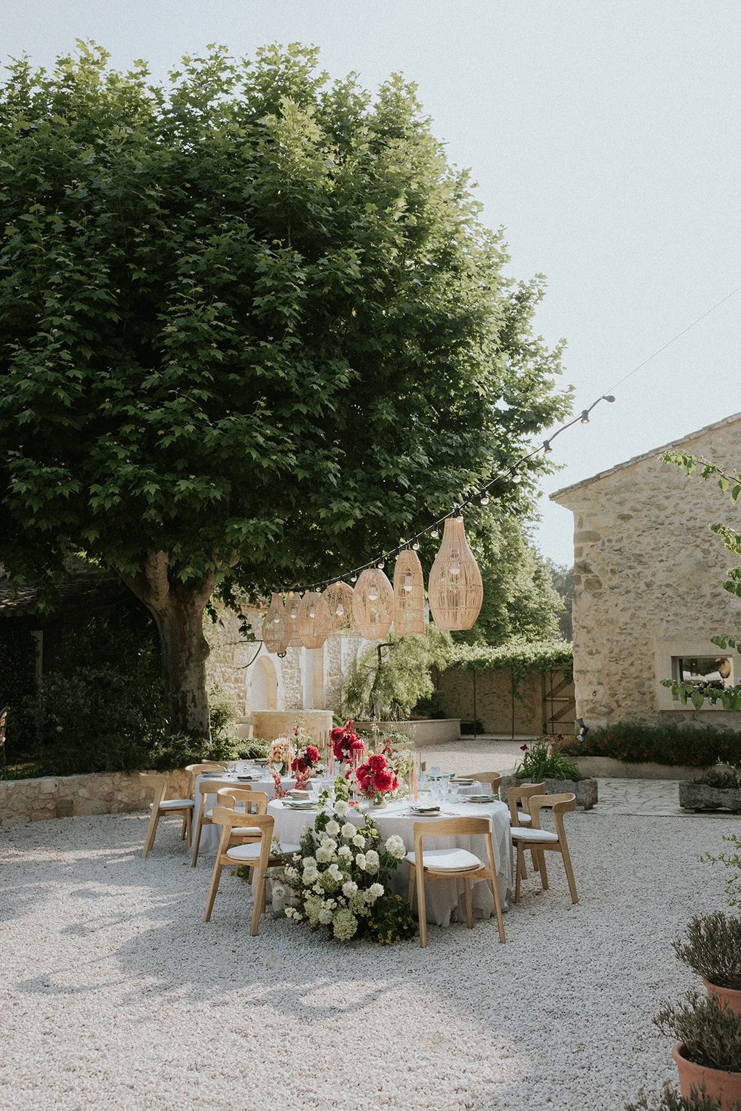 Outdoor wedding or event setup with a round table decorated with pink and white flowers, surrounded by wooden chairs, under a large leafy tree with hanging wicker lanterns and a stone building in the background.
