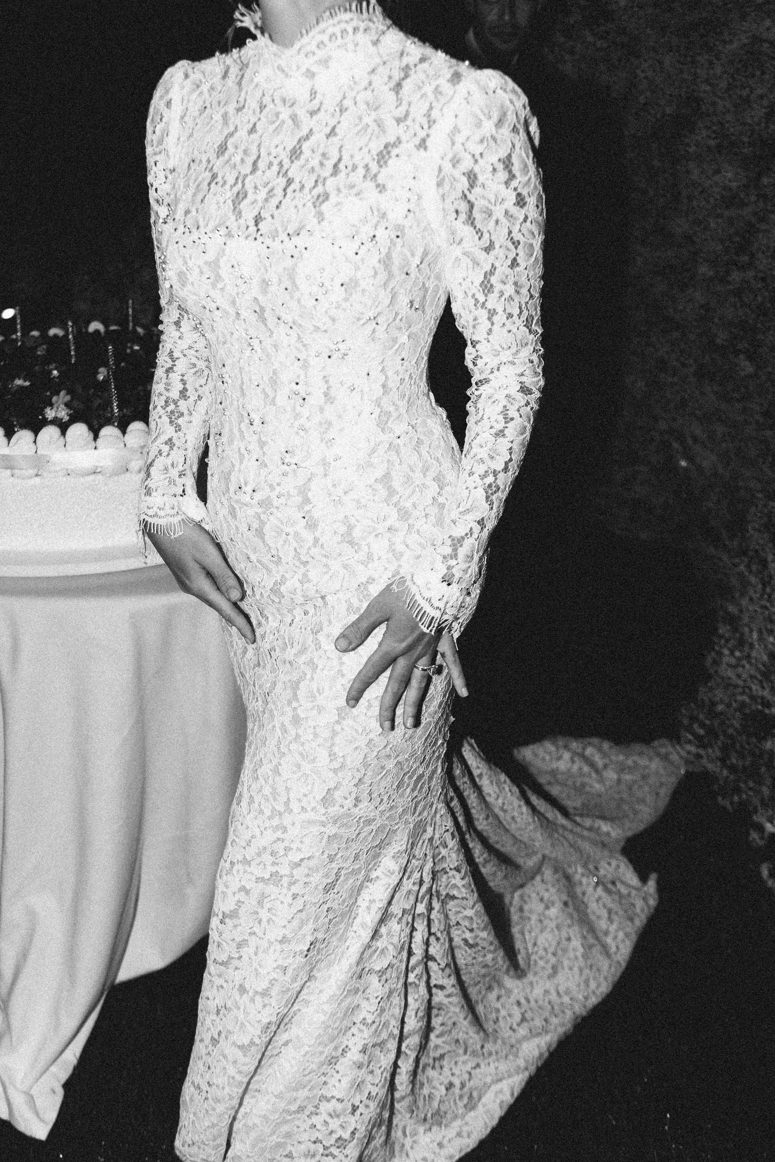 The bride wearing a long, white lace wedding dress with intricate floral patterns, standing with one hand on her hip and the other touching the side of the dress.