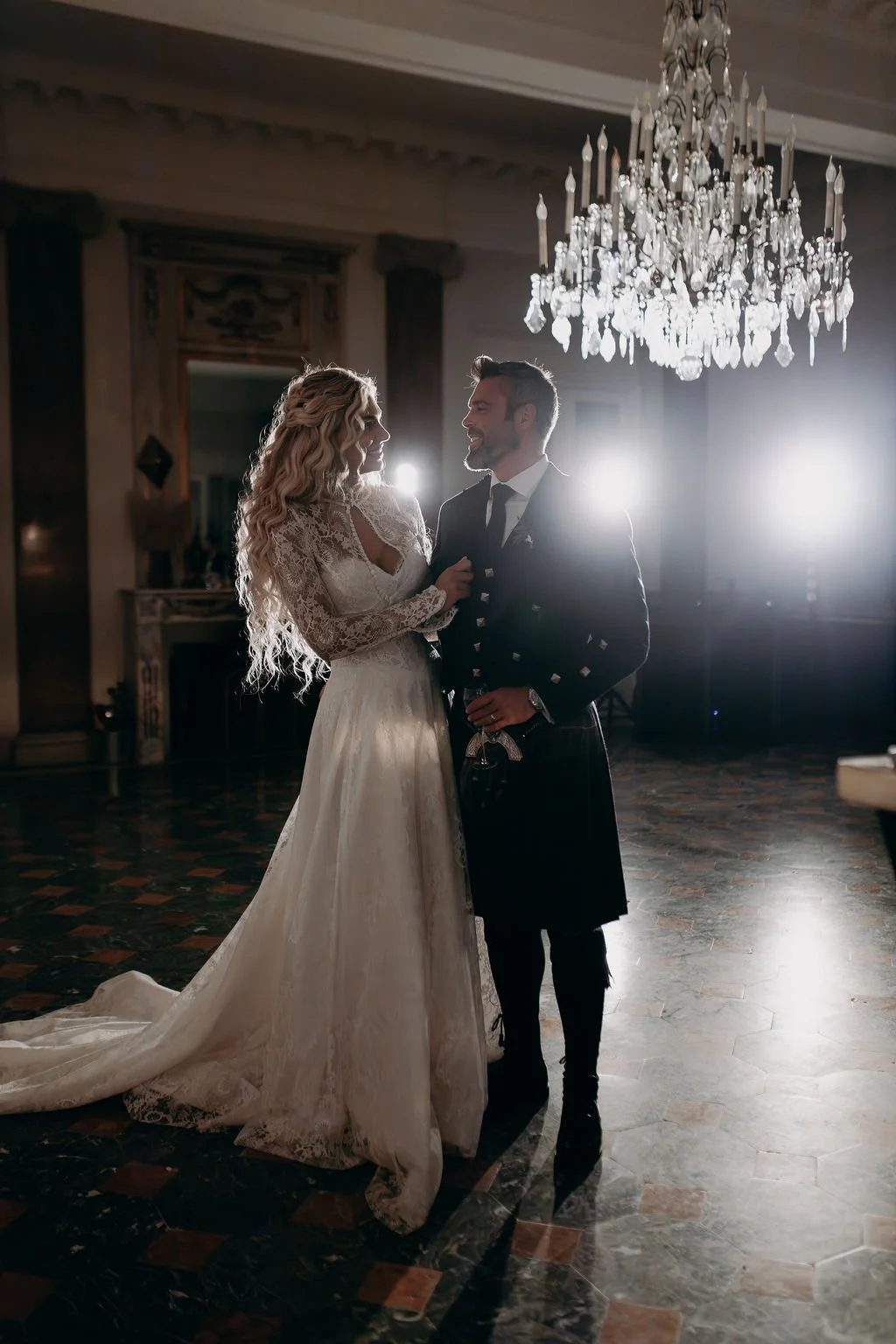 A bride and groom are dancing in a fancy hall with a chandelier overhead during their wedding.