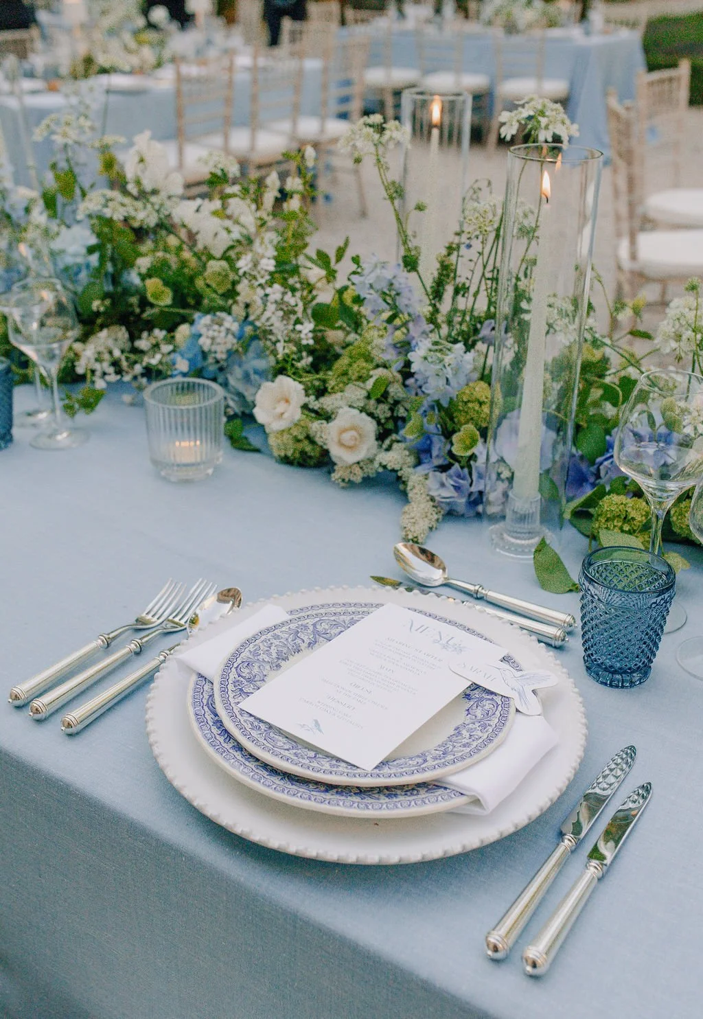 Elegant outdoor wedding table setting with floral centerpieces, candles, blue and white dinnerware, silver flatware, and a printed menu on top.