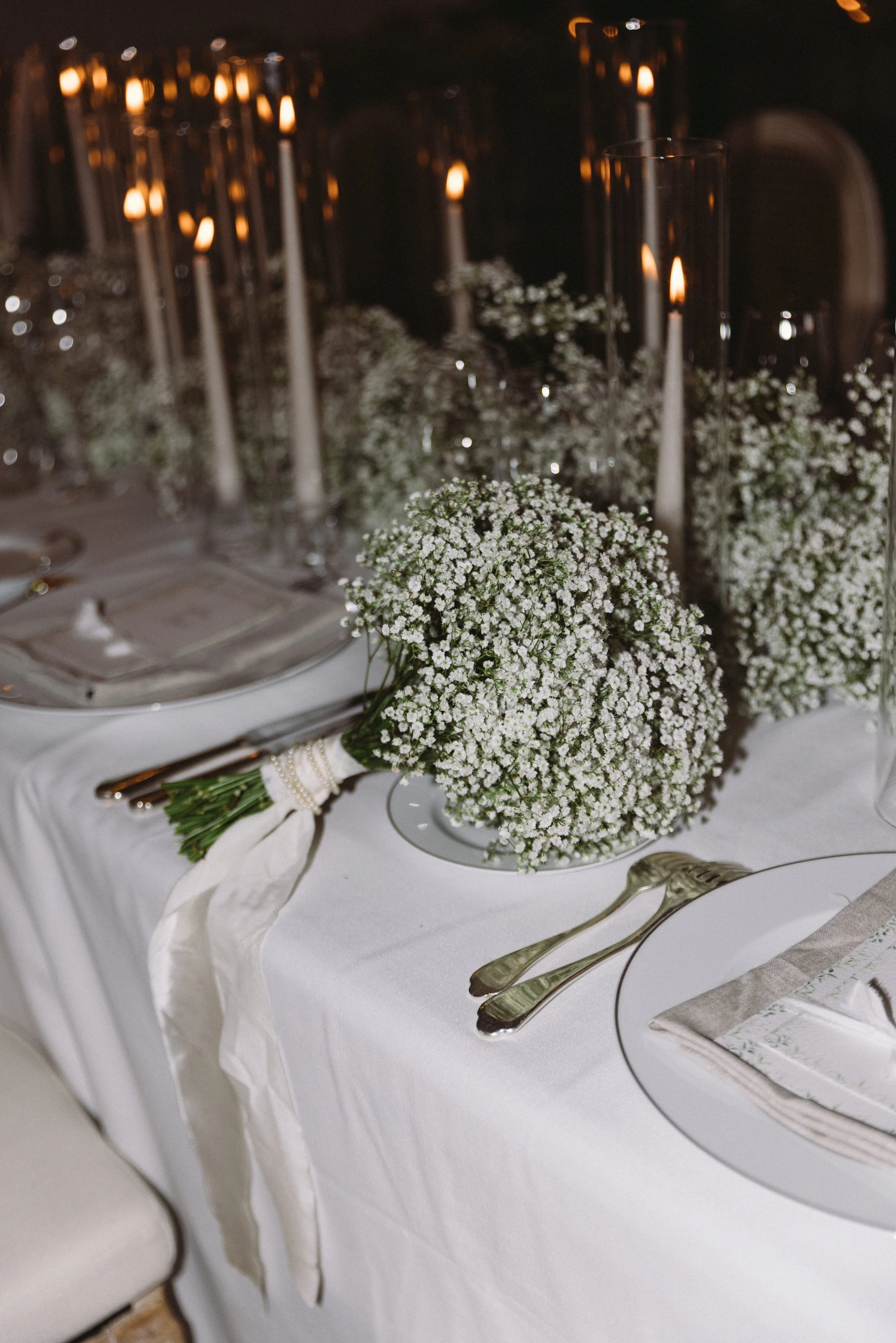 Elegant table setting with white tablecloth, floral centerpiece of baby's breath, tall candles in glass holders, and silverware.
