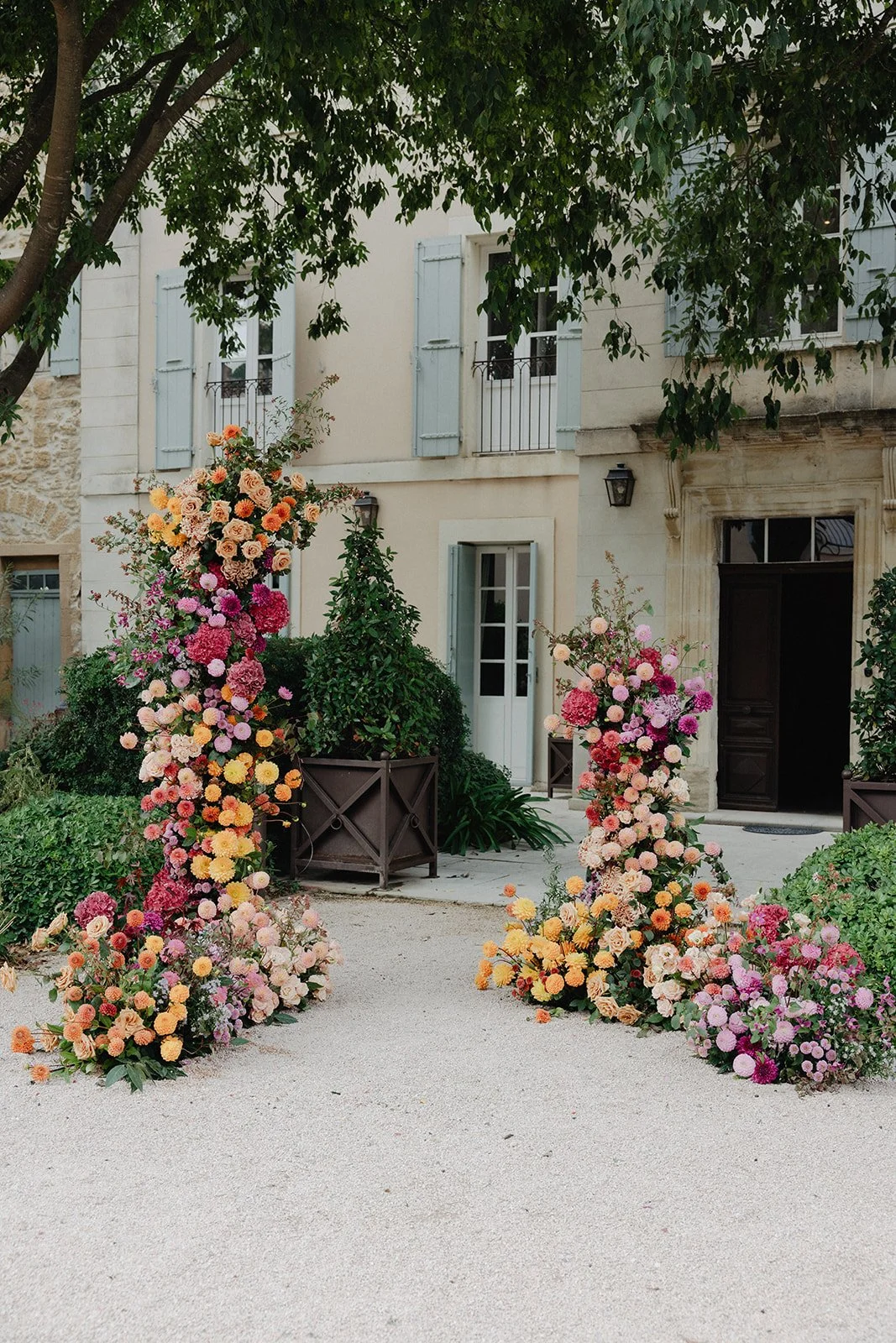 Decorative floral archway with colorful pink, yellow, peach, and purple flowers in front of a beige building with light blue shutters and a black door. Outdoor ceremony in Provence during fall season.