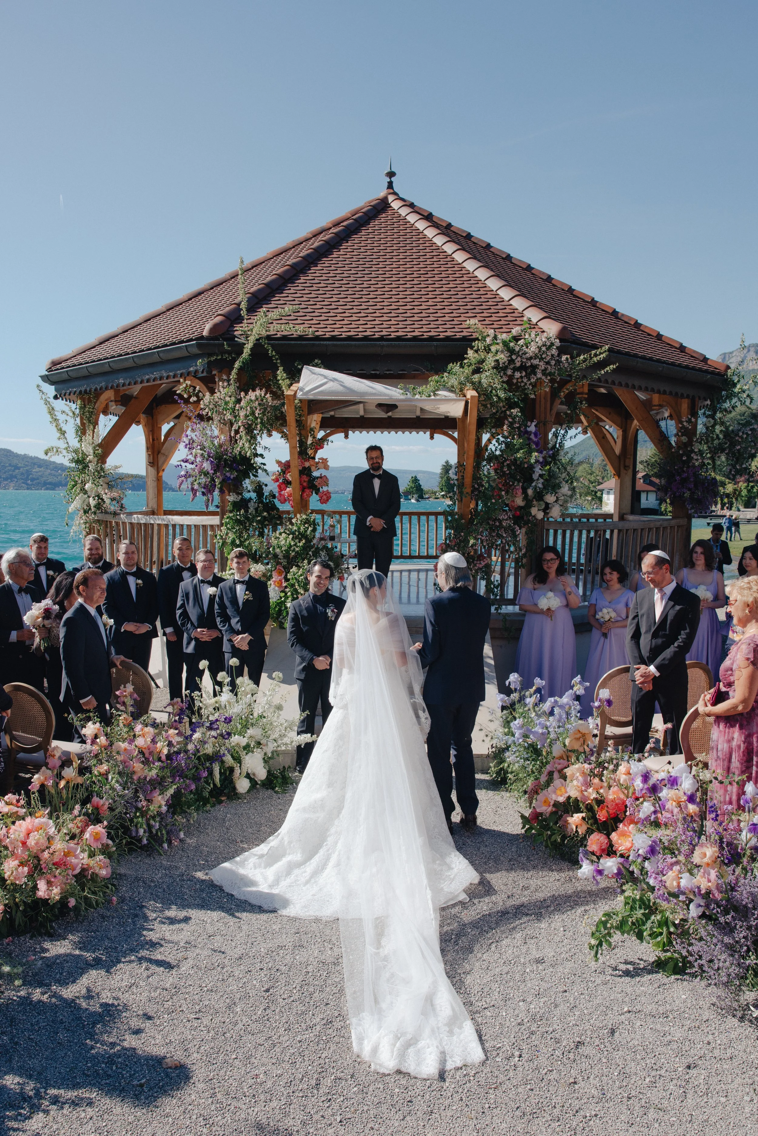 A wedding ceremony outdoors by a lake with a wooden pavilion at Palace de Menthon, bride and groom standing with an officiant, surrounded by floral arrangements, guests, and wedding party.