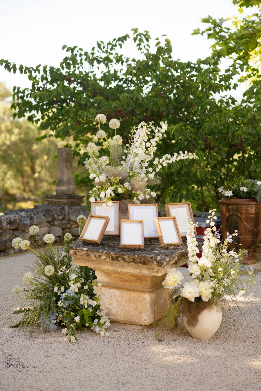A seating chart display with framed photos, white flowers, and greenery on a stone pedestal, set against a stone wall and lush trees.