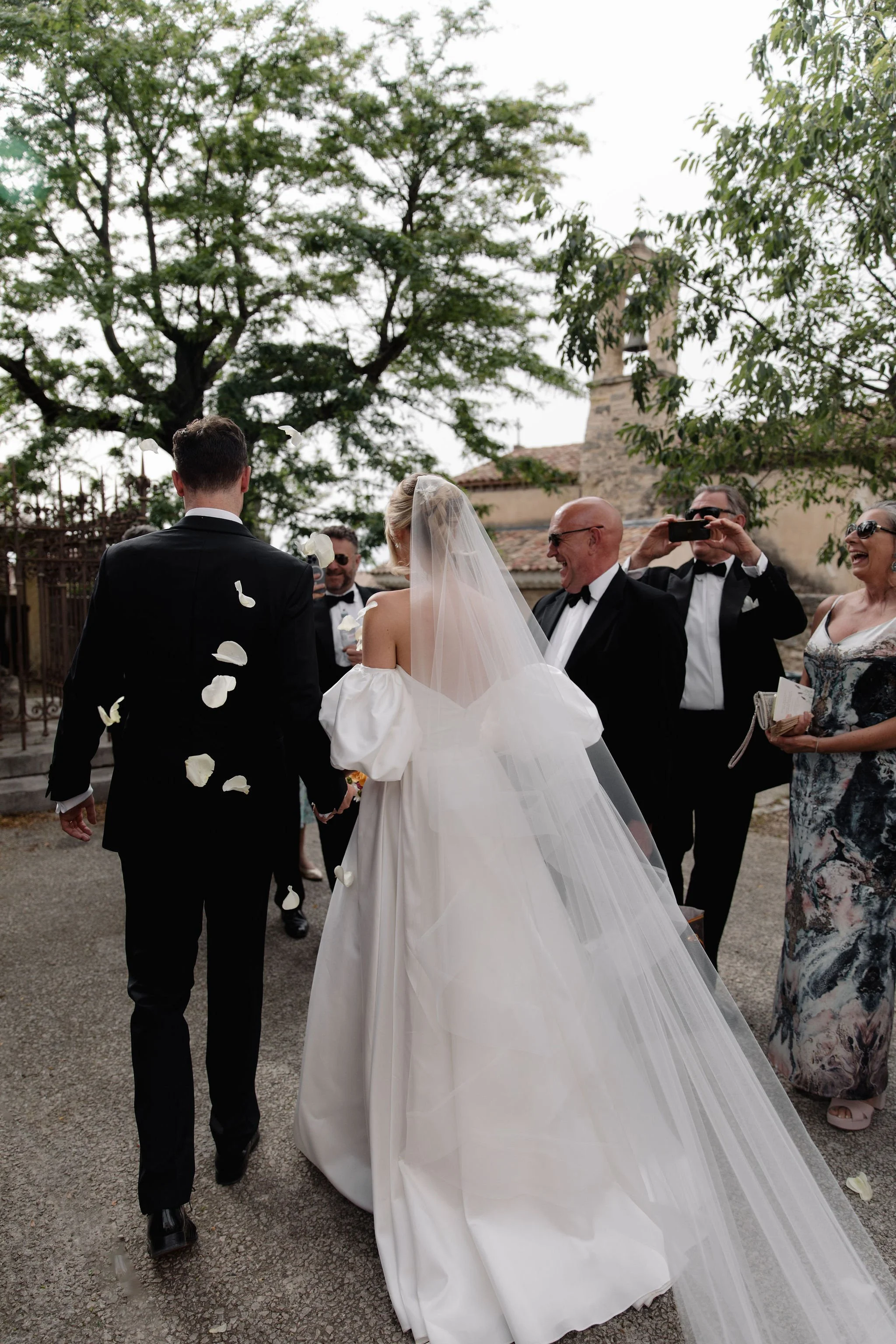 Bride and groom walking outdoors with guests throwing flower petals, surrounded by trees and a stone building.