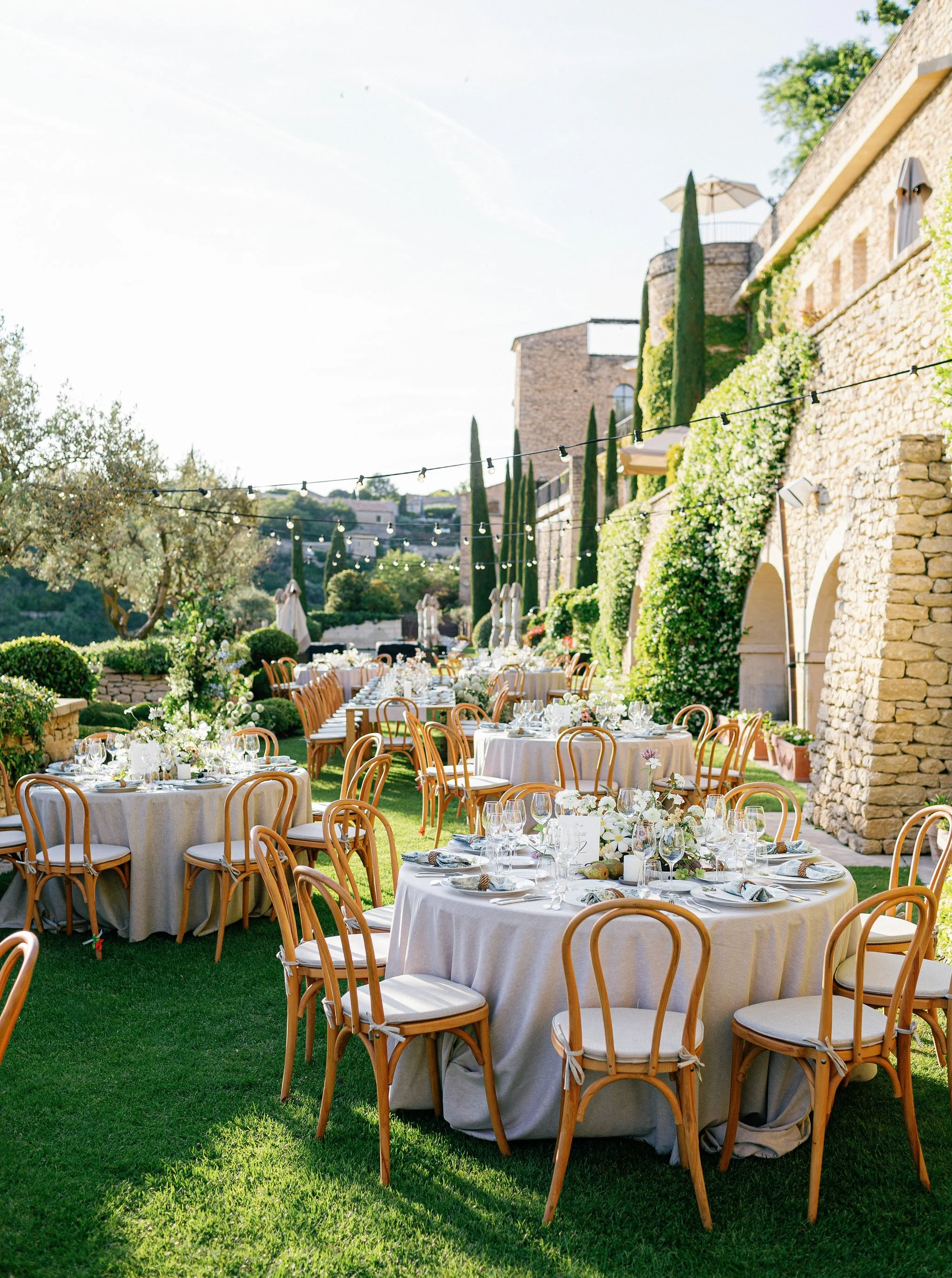 Outdoor wedding reception with round tables covered in white tablecloths, set with glasses, plates, and floral centerpieces, arranged on a lush green lawn with a stone building and string lights overhead.