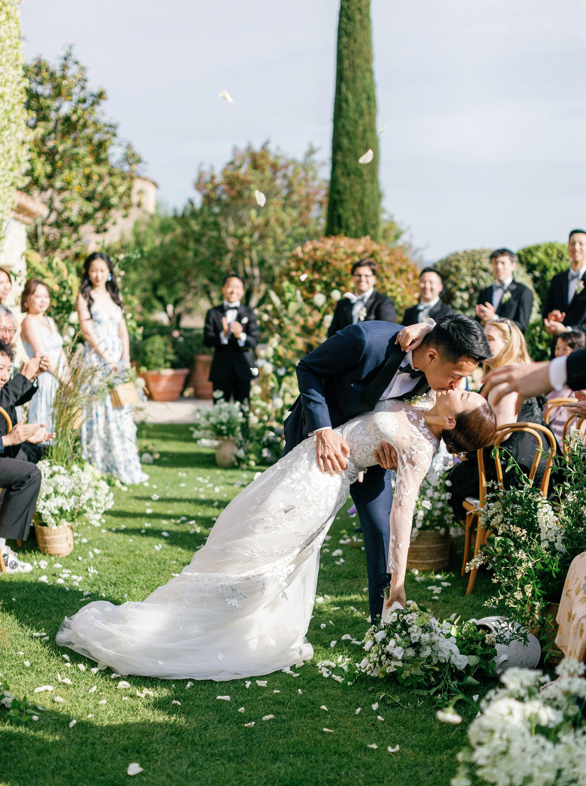 A wedding ceremony outdoors with a bride and groom sharing a deep kiss, surrounded by seated guests and lush greenery.