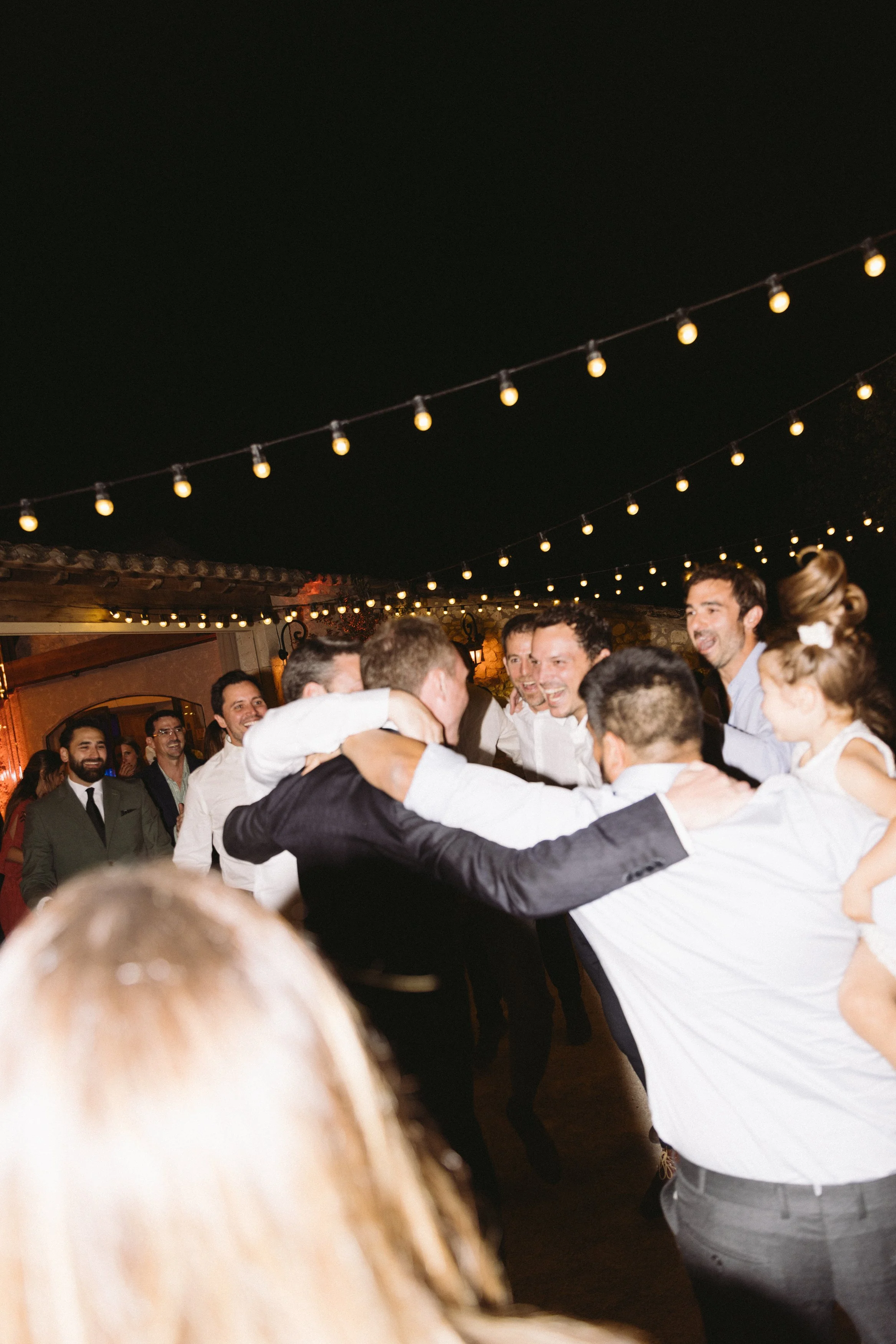Group of people dancing and celebrating at a party with string lights overhead at night.