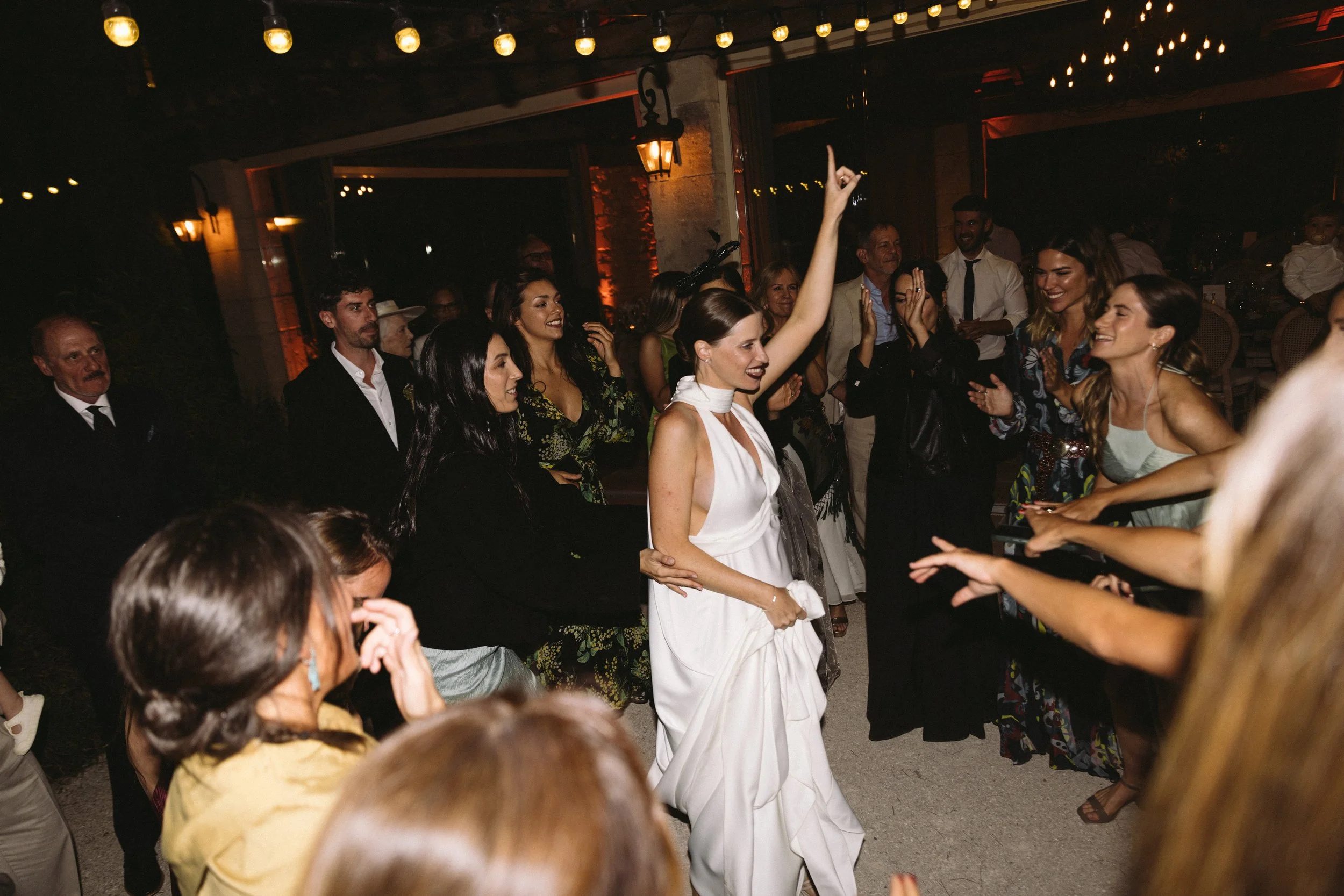 The bride in a white dress dancing and raising her arm at a lively party with surrounded smiling guests at night.