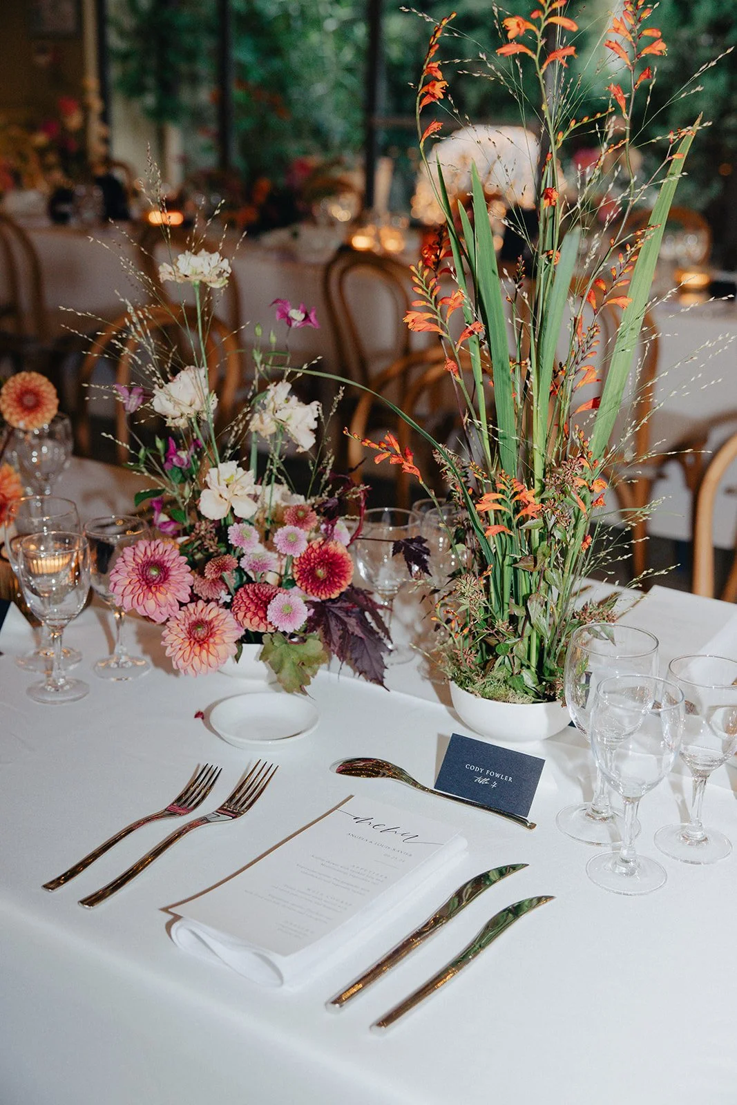 Table setting with floral centerpieces, including pink, white, and purple flowers, surrounded by empty wine glasses and cutlery, at a formal event.