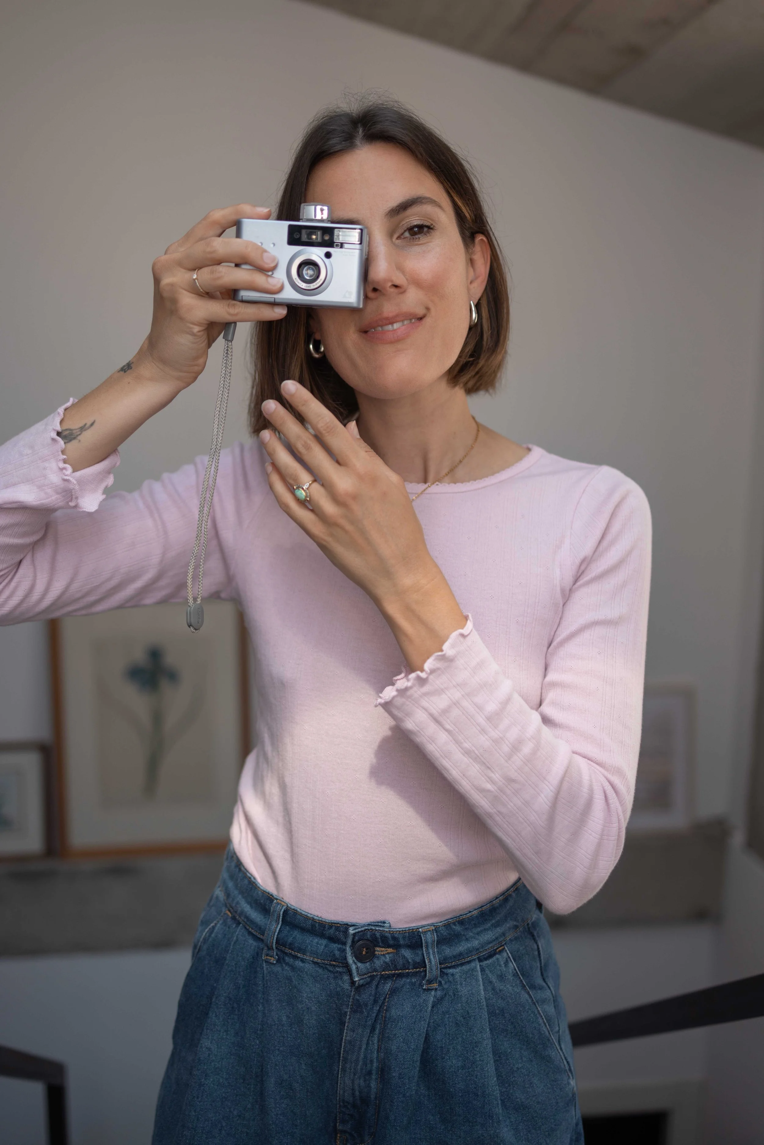 Jeune femme souriante tenant un appareil photo devant son visage, portant un haut rose et une jupe en jean, dans un intérieur.
