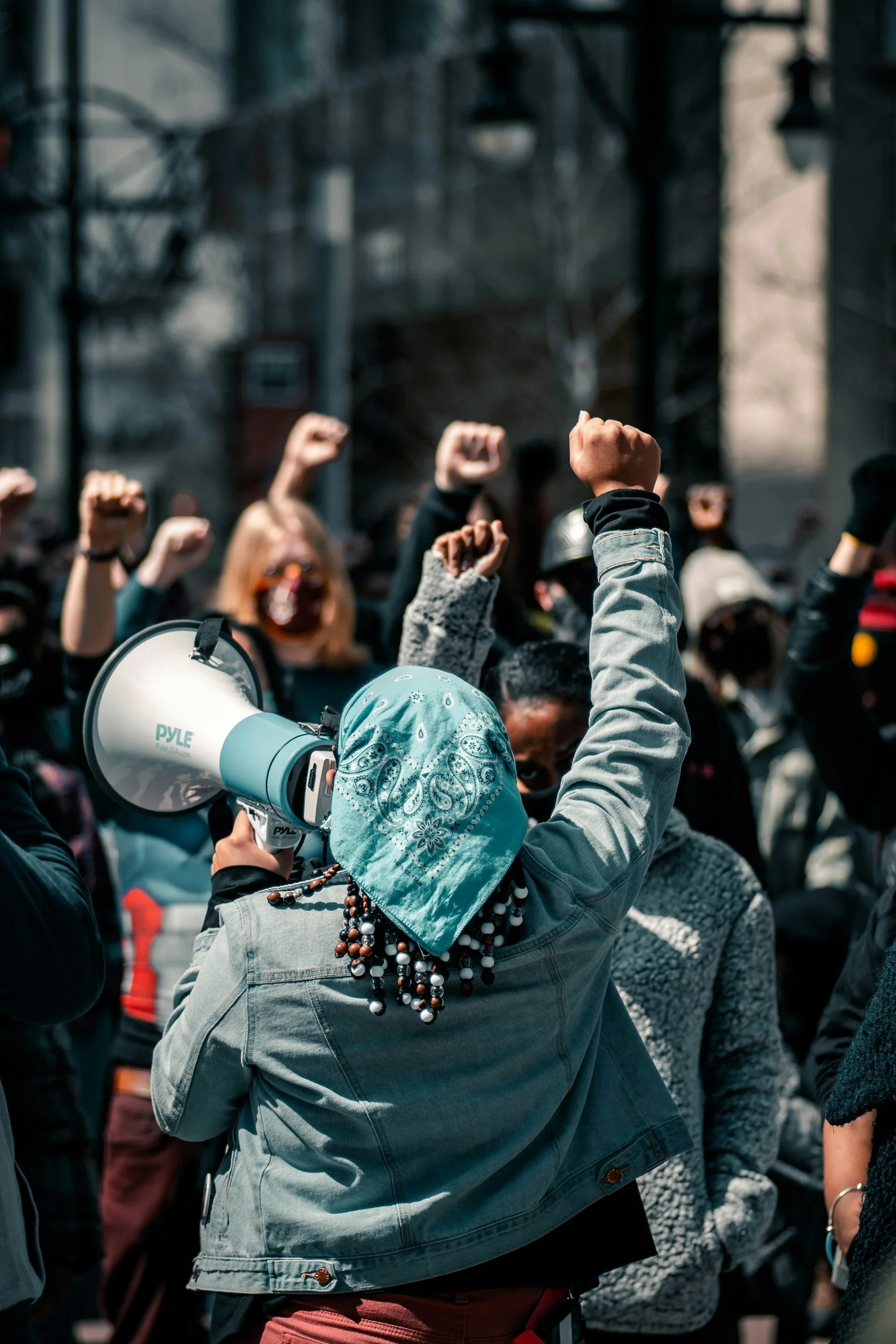 A Black woman faces a crowd, her back to the camera. One hand is held in the air in a fist, and in her other hand is a megaphone. She is speaking to the crowd.